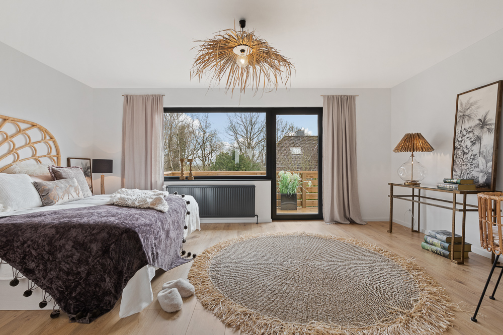 Helles Schlafzimmer mit großem Fenster, Doppelbett, rundem Teppich und Schreibtisch aus Holz.