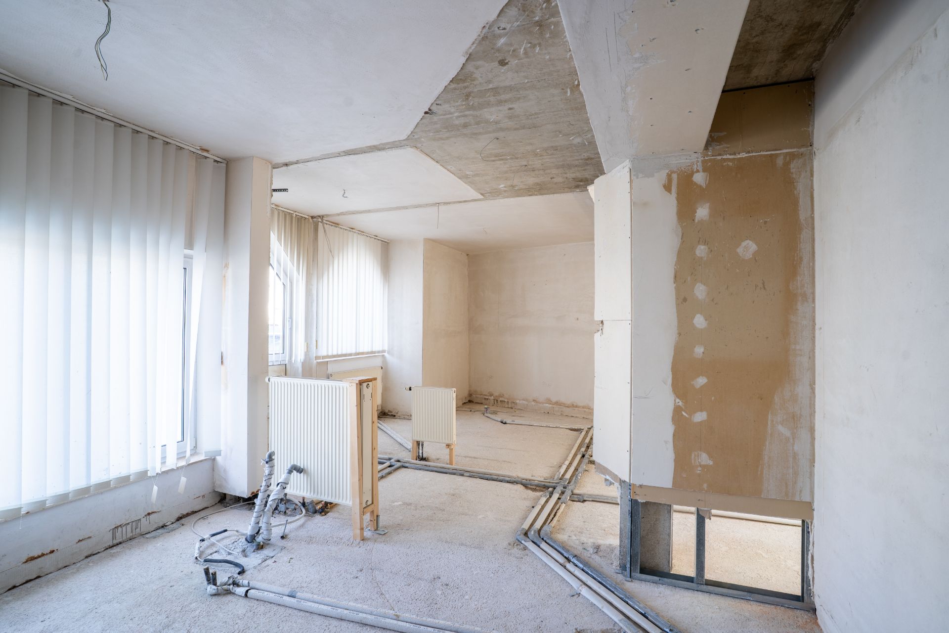 Heller Raum in Renovierung mit freiliegenden Rohren, Heizkörpern und großen Fenstern mit Jalousien.