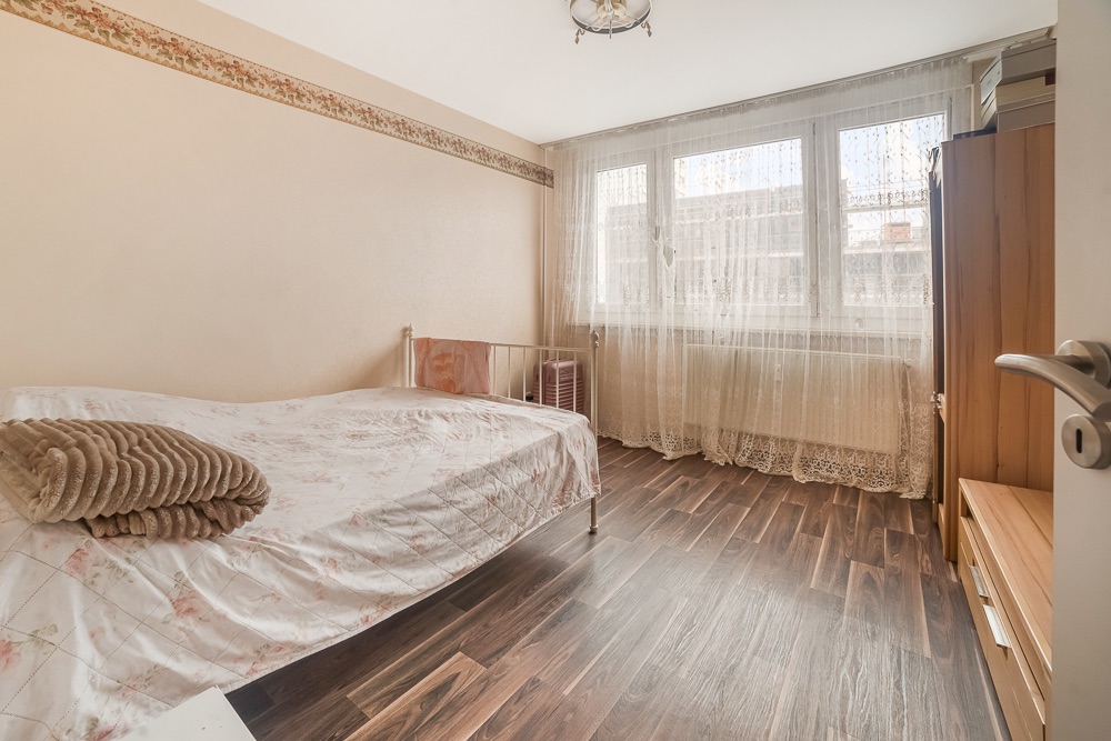 Helles Schlafzimmer mit Einzelbett, Holzboden, Gardinen vor großen Fenstern und Schrank.