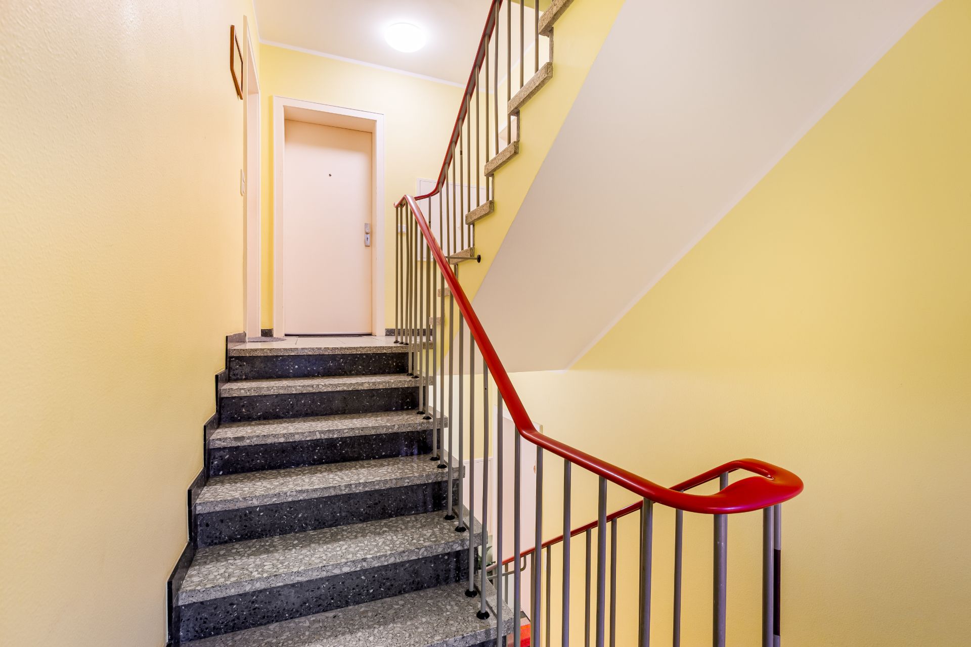 Treppenhaus mit grauer Treppe, gelben Wänden und rotem Handlauf in einem Wohngebäude.