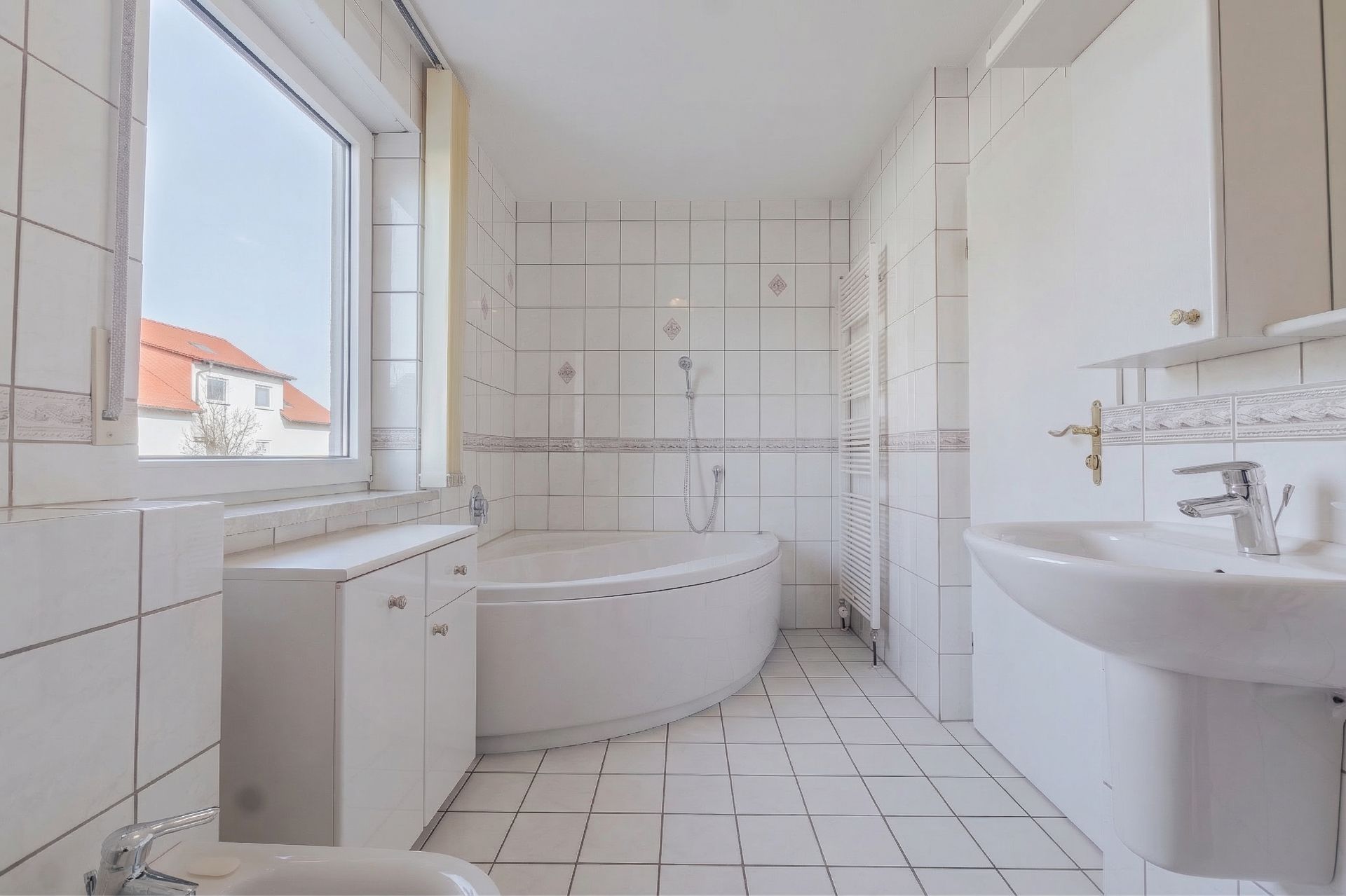 Helles Badezimmer mit Eckbadewanne, Waschbecken, Fenster und weißen Fliesen.