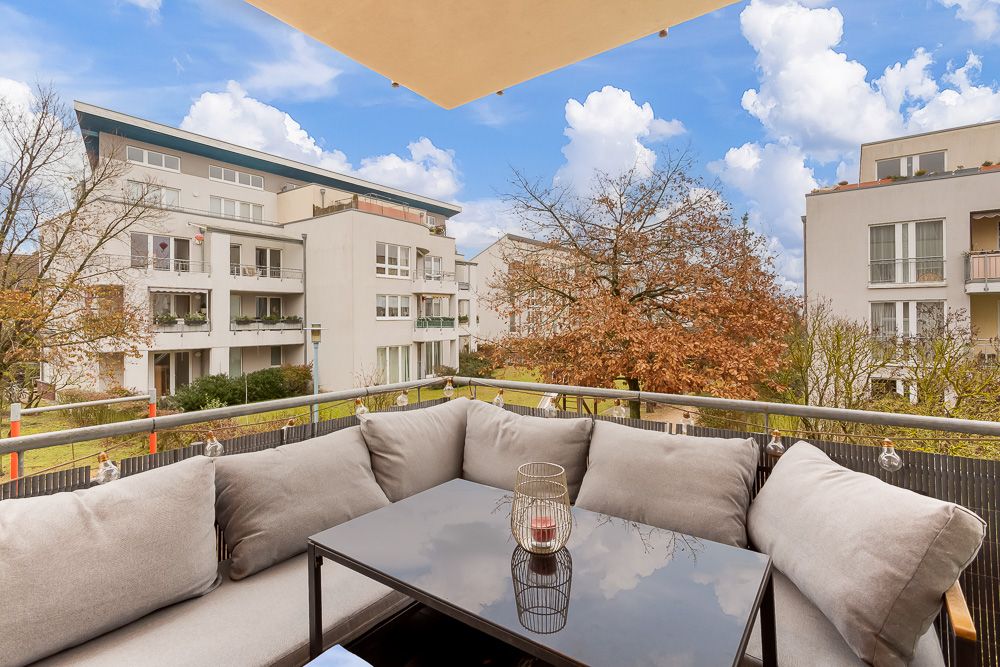 Balkon mit grauem Sofa, Glastisch und Blick auf moderne Wohngebäude und Bäume.