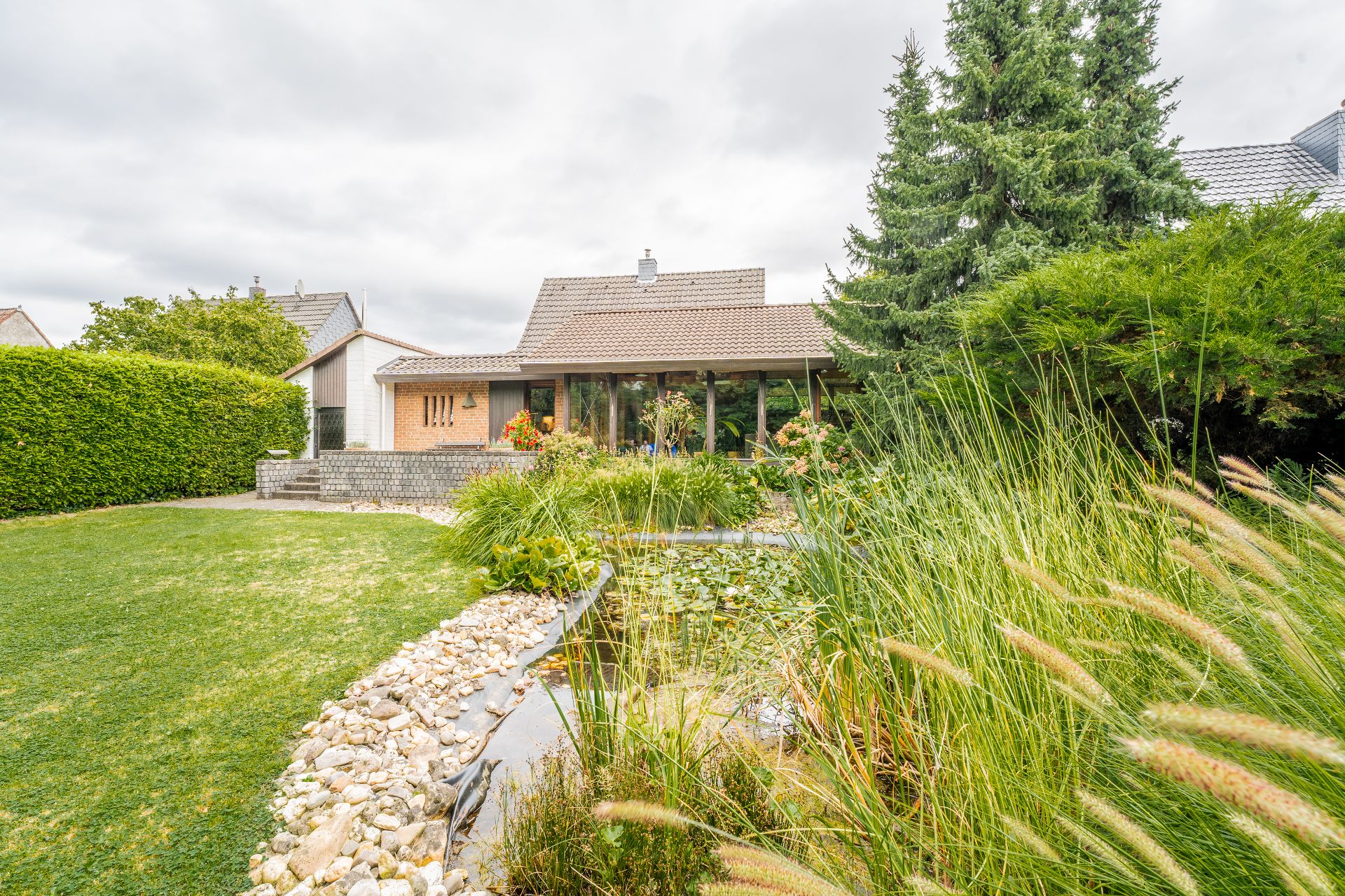Haus mit großem Garten, Teich, viel Grün und Bäumen, aufgenommen bei bewölktem Himmel.