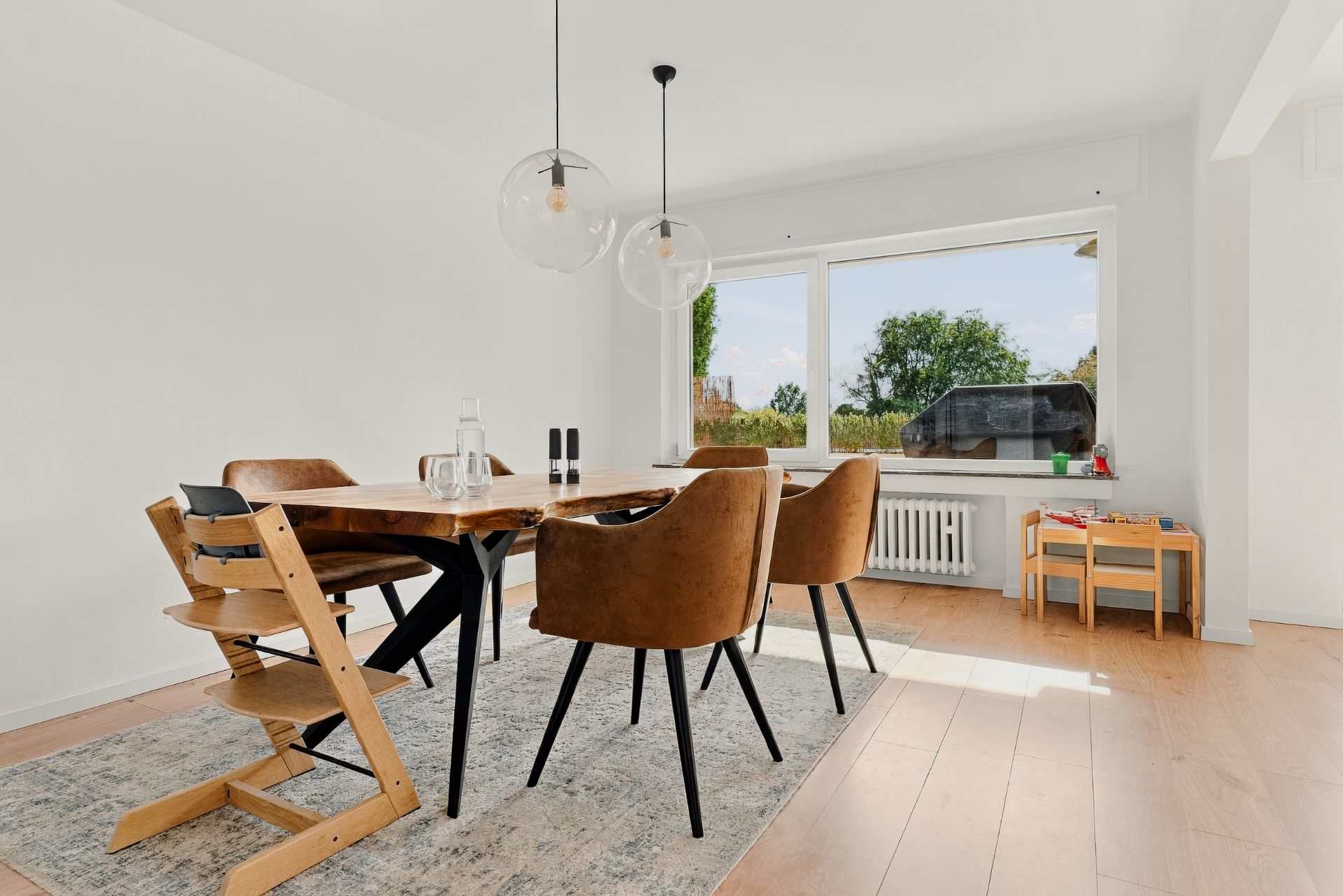 Esszimmer mit Holztisch, vier Stühlen, Kinderstuhl und großem Fenster mit Blick ins Grüne.