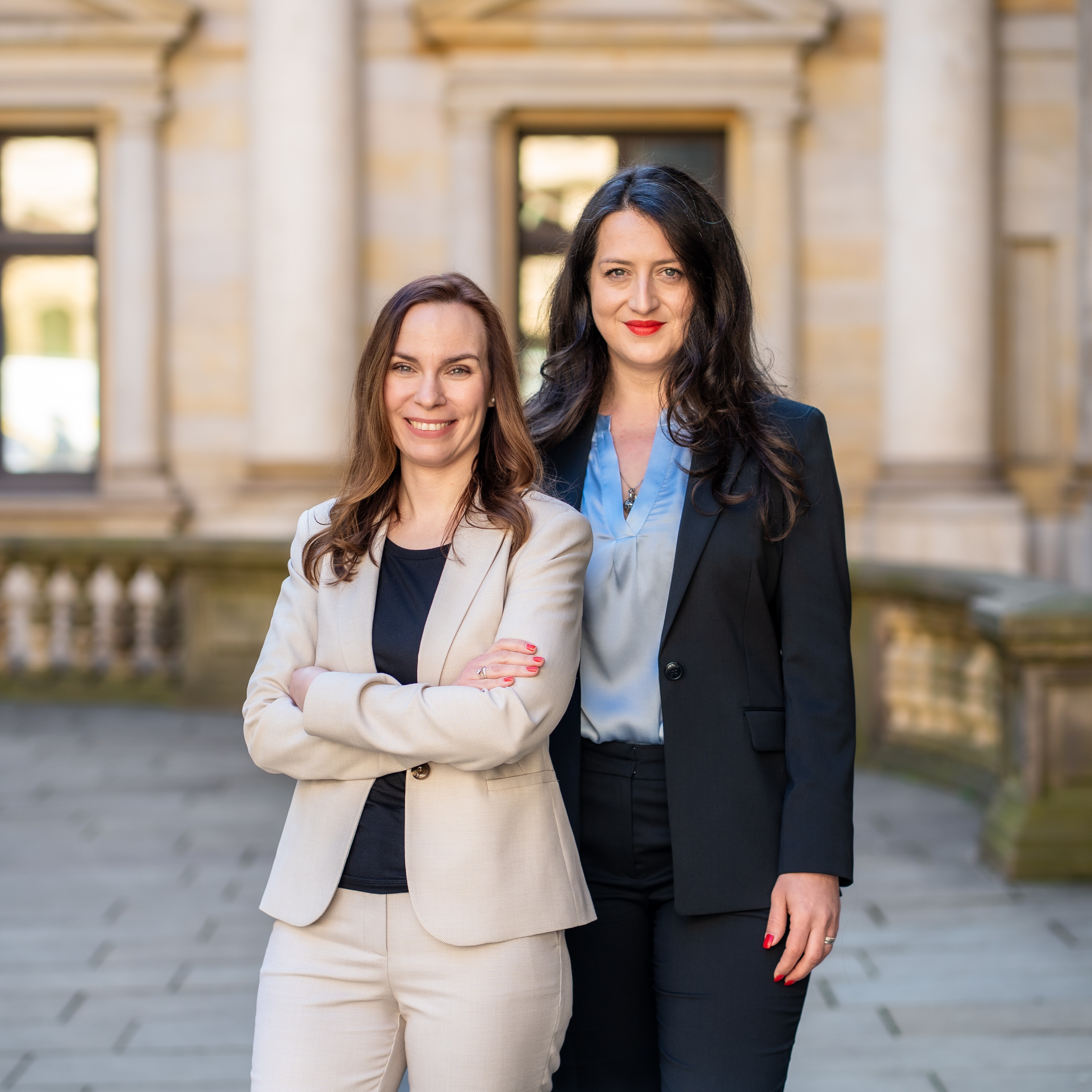 Zwei Frauen in Businesskleidung stehen draußen vor einem historischen Gebäude.