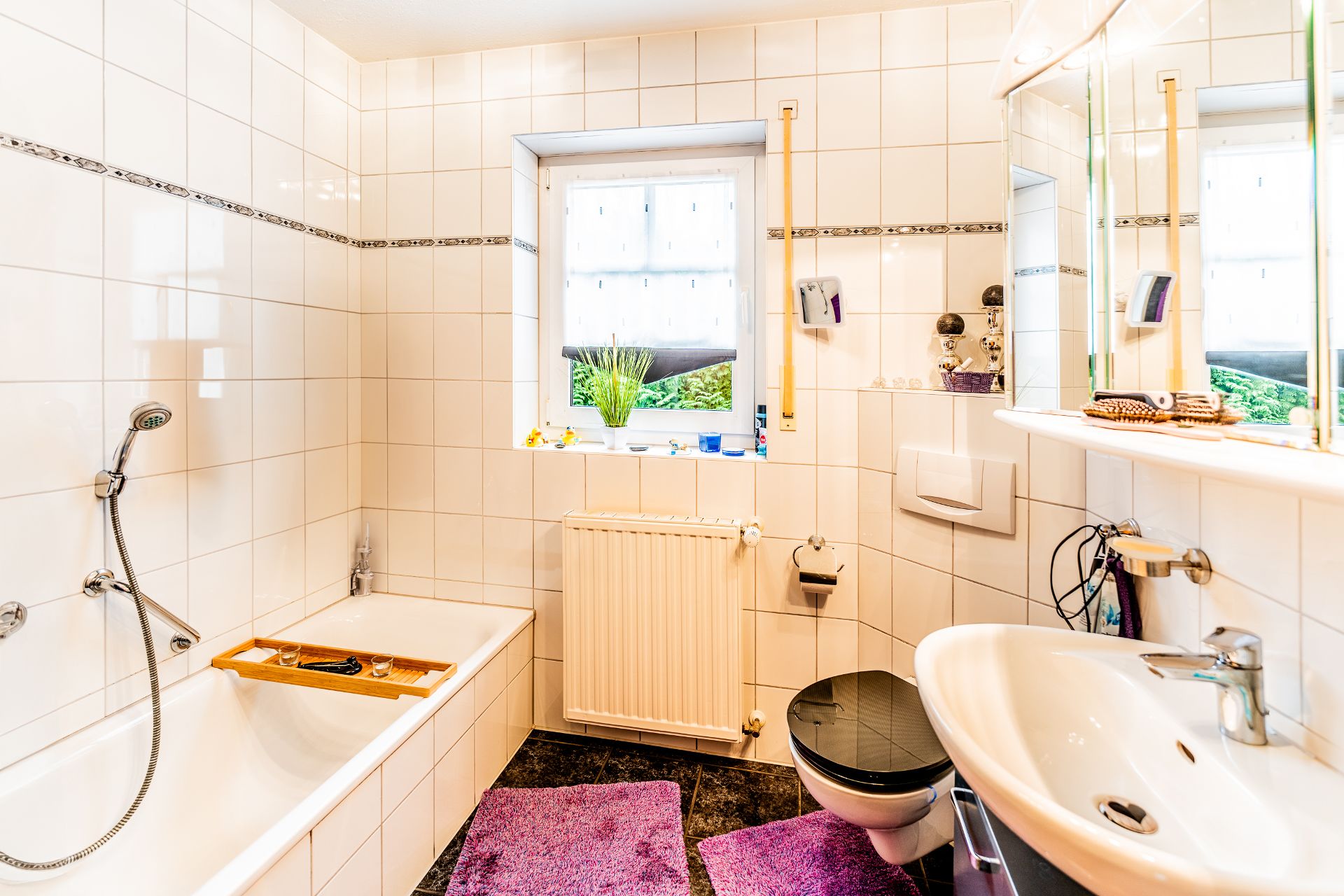 Helles Badezimmer mit Badewanne, Waschbecken, WC, Fenster und lila Teppichen.