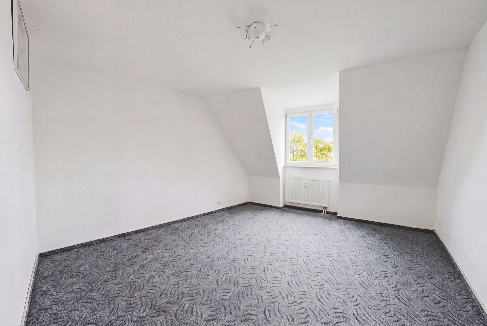 Leeres Zimmer mit grauem Teppichboden, weiß gestrichenen Wänden und einem Fenster mit Blick ins Grüne.