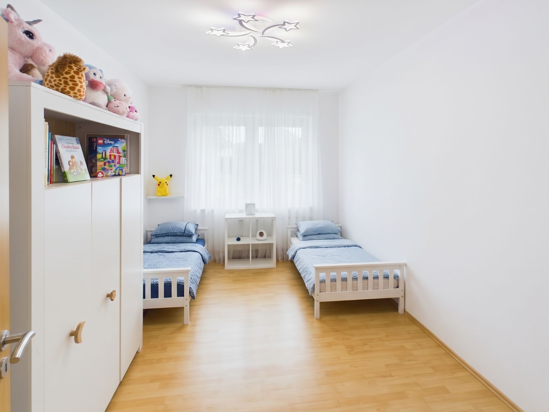 Helles Kinderzimmer mit zwei Einzelbetten, Holzfußboden und einem weißen Schrank mit Kuscheltieren.