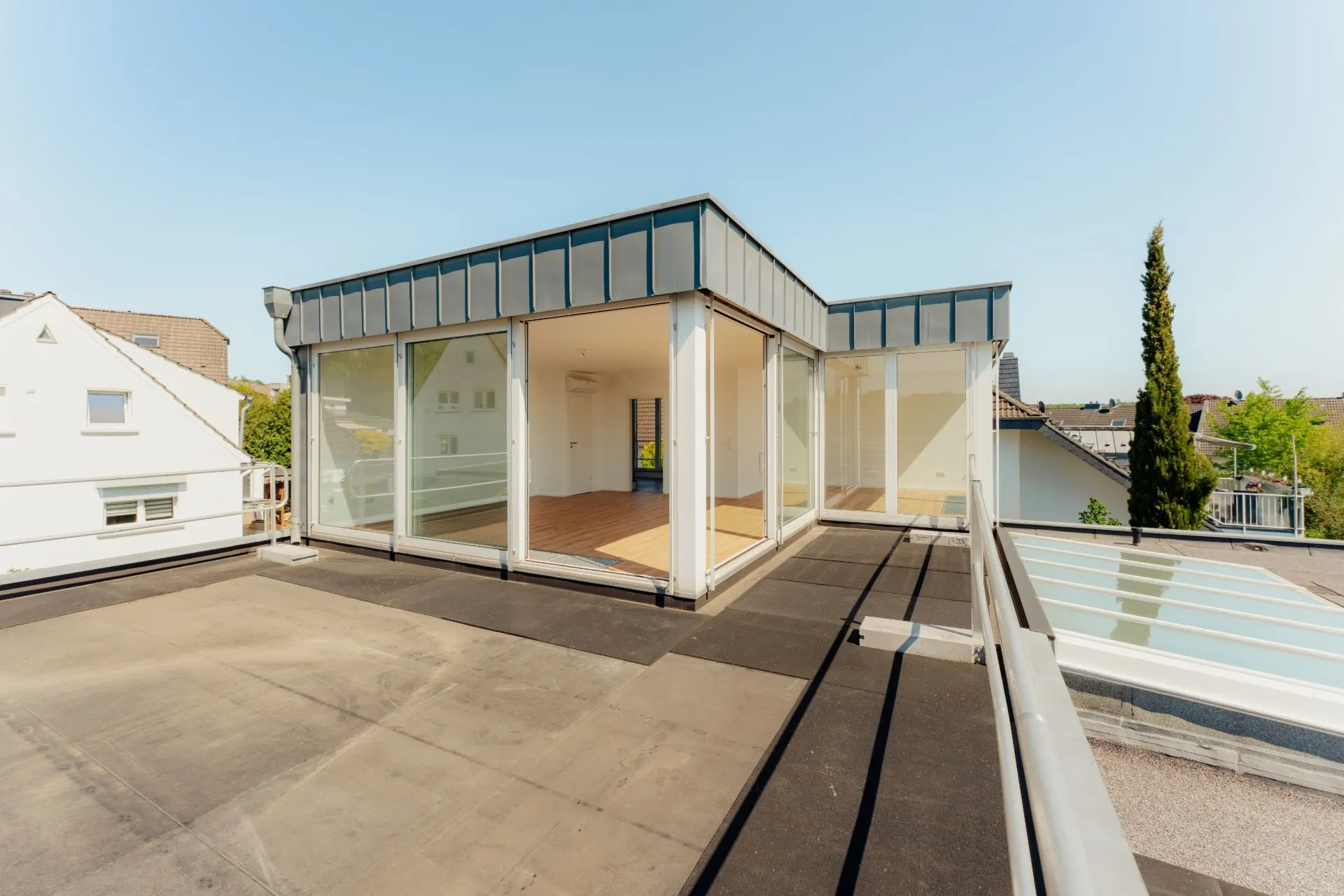 Moderne Dachterrasse mit Glaswänden und Blick auf Nachbarhäuser und Bäume.