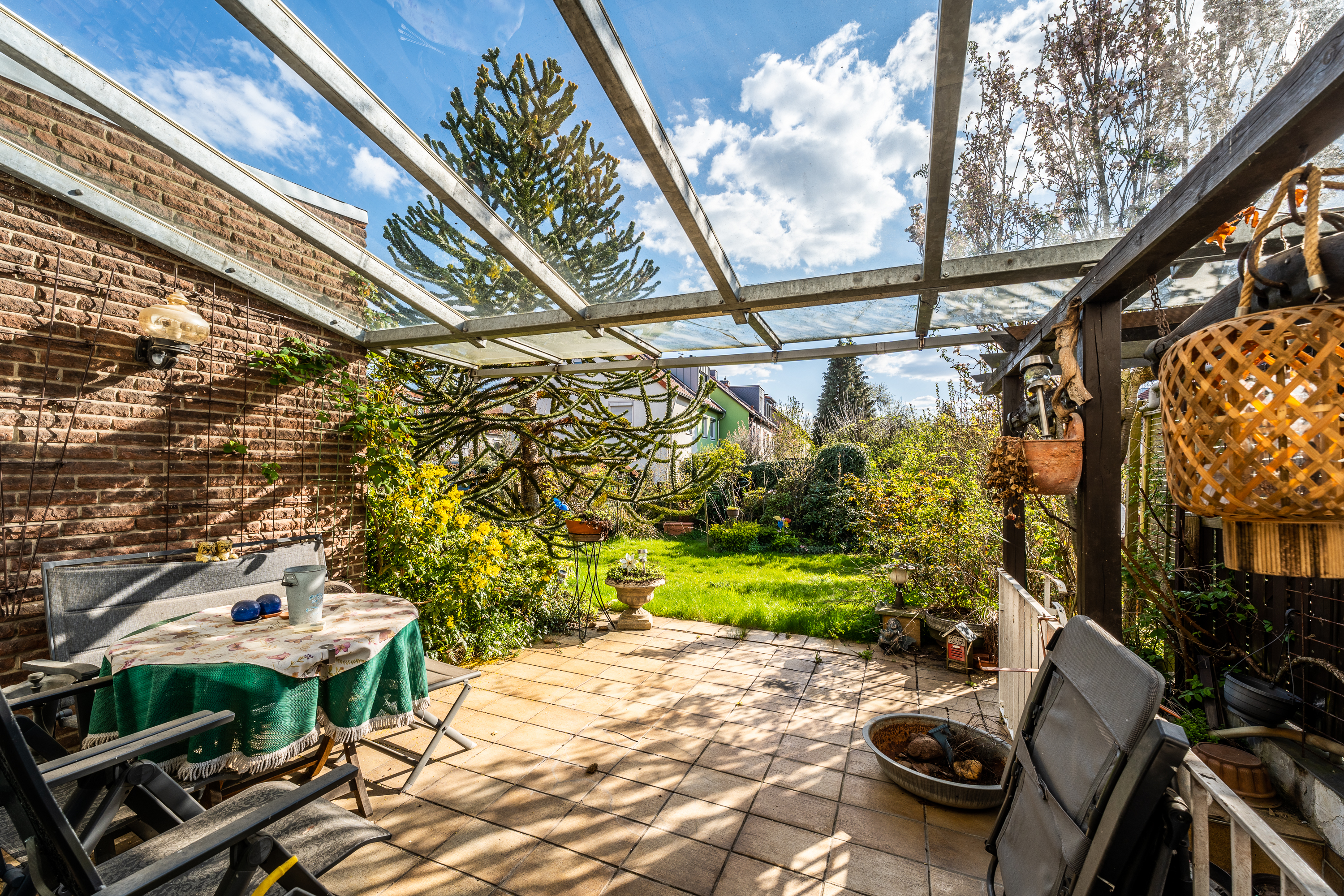 Überdachte Terrasse mit Tisch, Stühlen und Blick auf gepflegten Garten mit vielen Pflanzen.