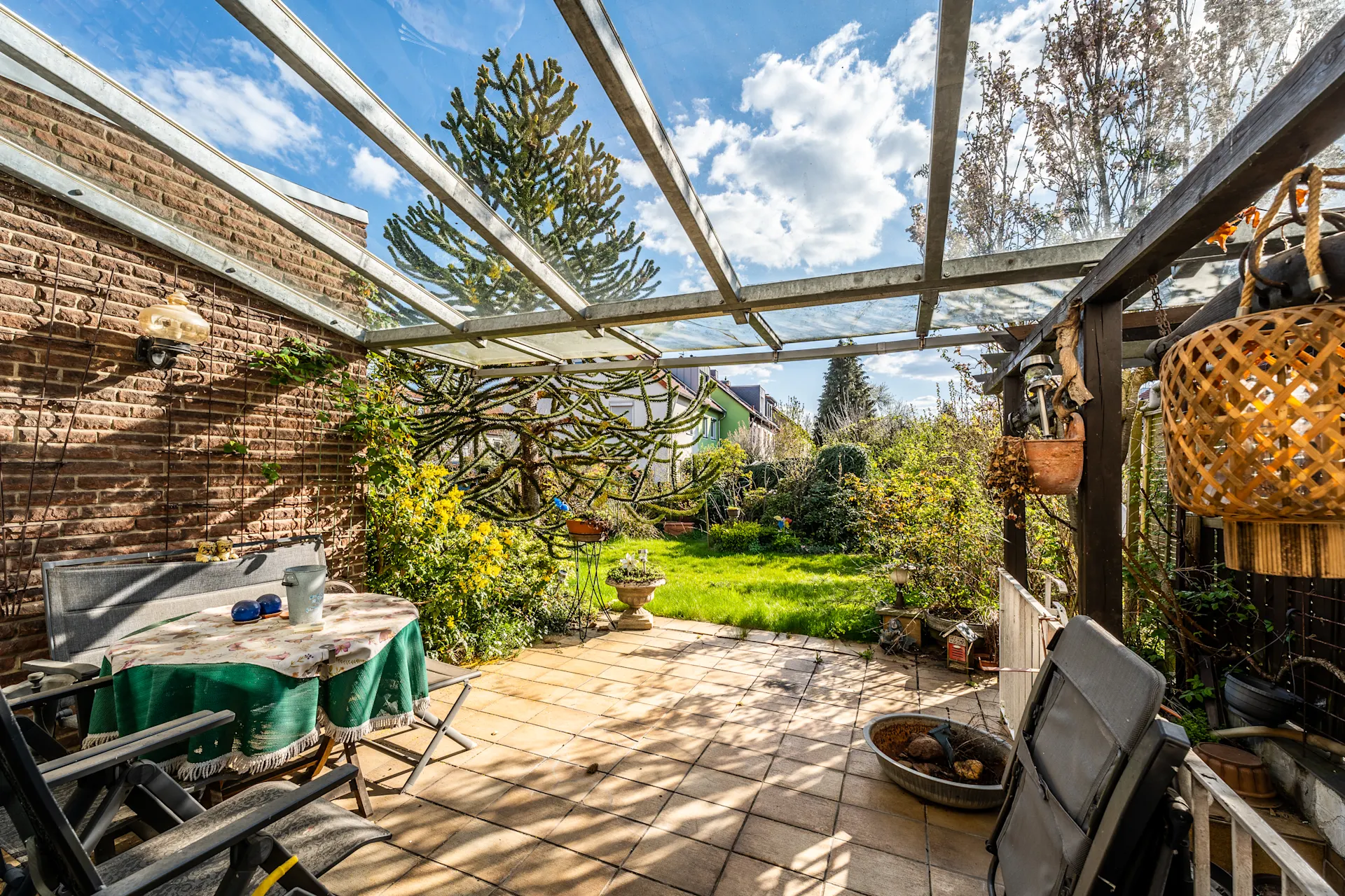 Überdachte Terrasse mit Tisch, Stühlen und Blick auf gepflegten Garten mit vielen Pflanzen.