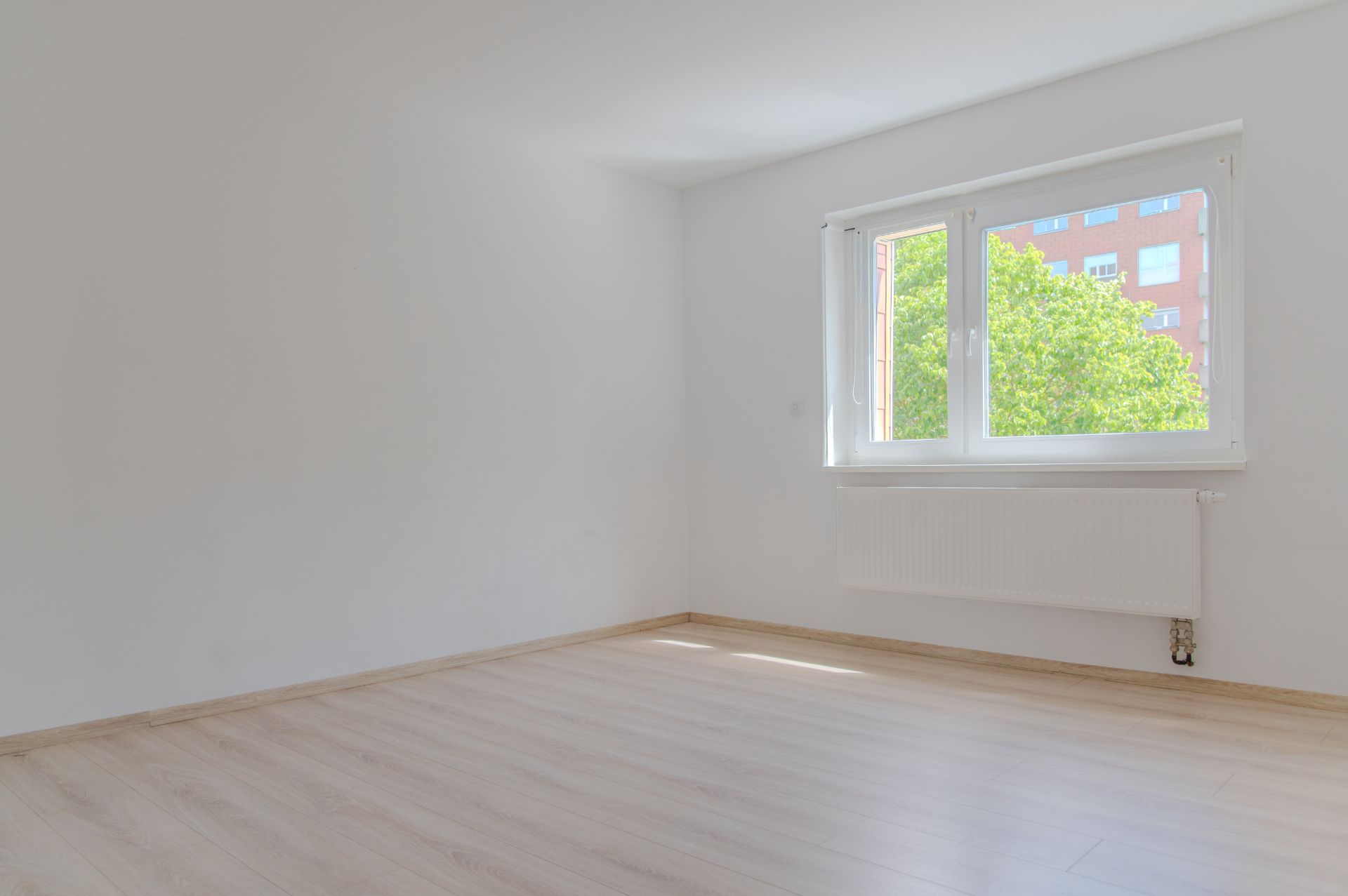 Heller, leerer Raum mit Holzfußboden und großem Fenster mit Blick auf Bäume.