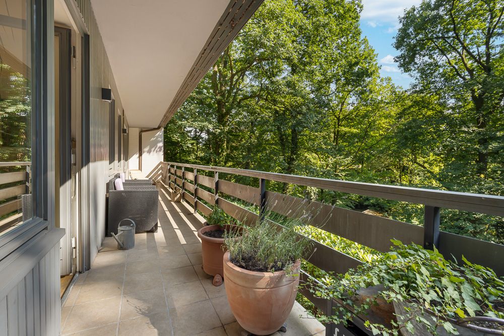 Langer Balkon mit Pflanzen in Töpfen, Sitzmöbeln und Blick auf grüne Bäume.