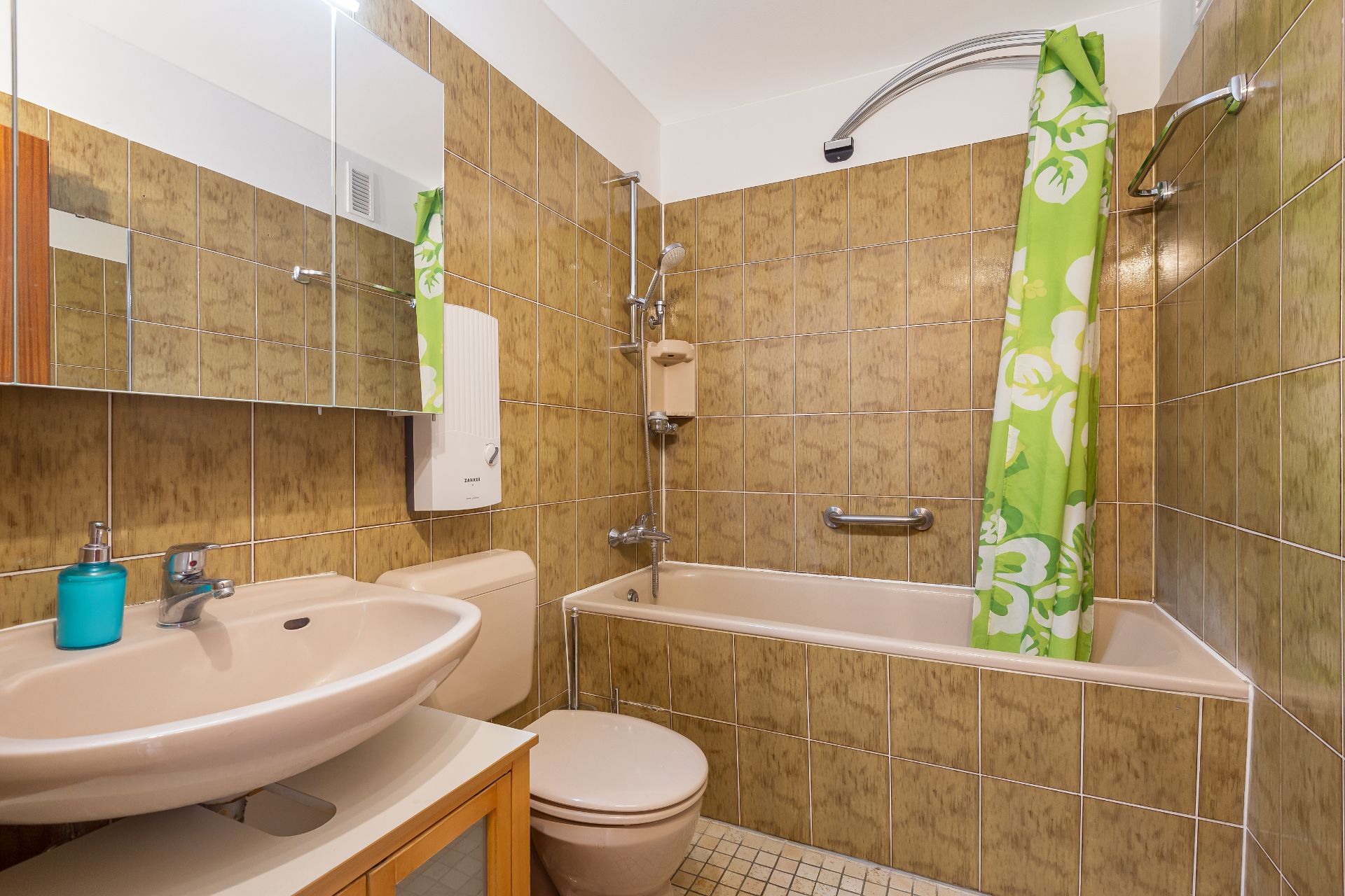 Badezimmer mit braunen Fliesen, Badewanne, grünem Duschvorhang, Waschbecken und Spiegel.