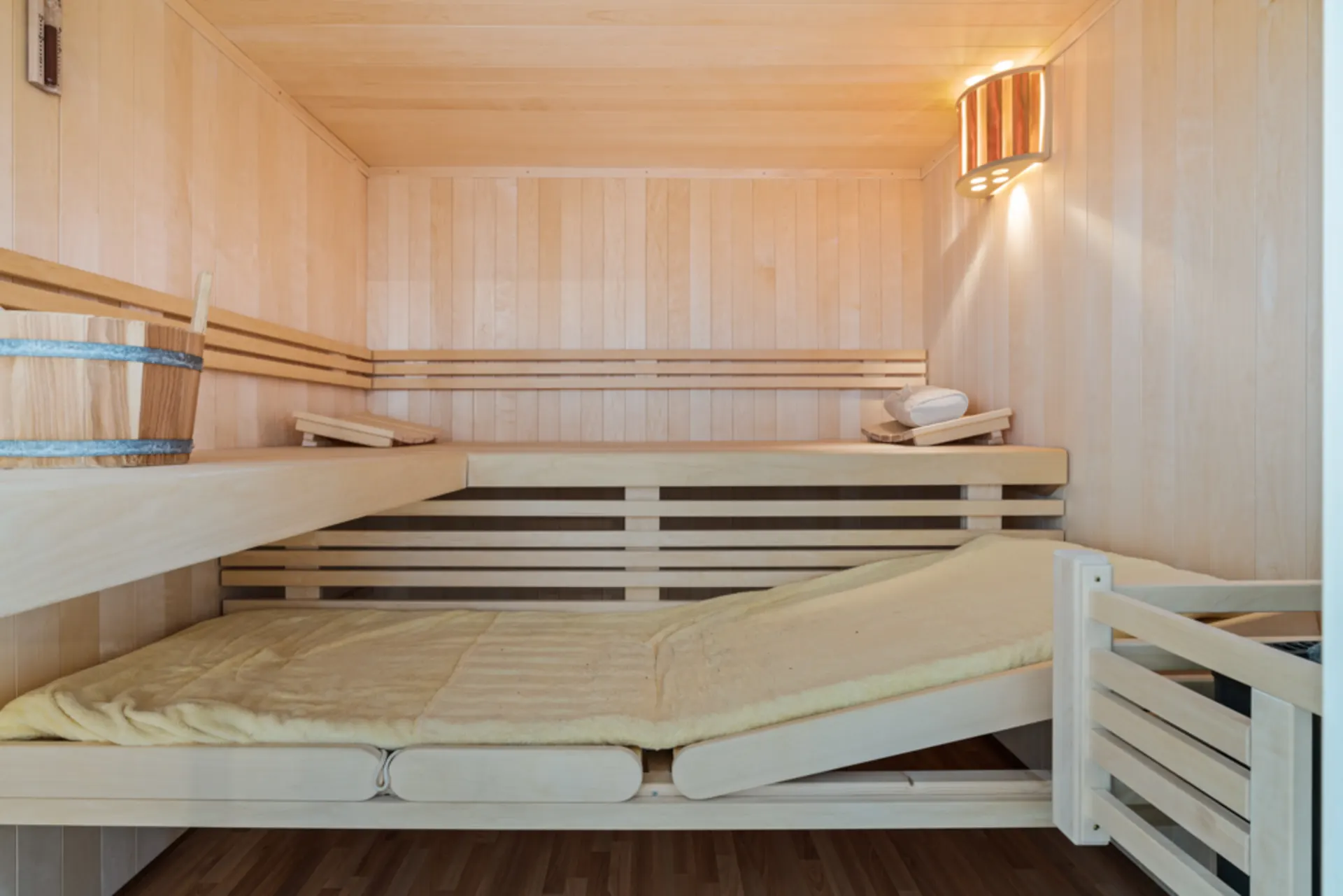 Helle Holzsauna mit Liegebank, Eimer und Kelle, an der Wand eine Lampe.
