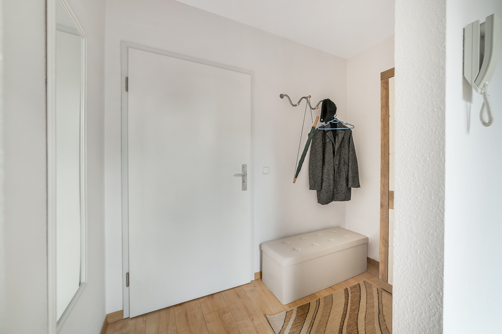 Heller Flur mit weißer Tür, Garderobe, Mantel, Regenschirm, Sitzbank und Spiegel.