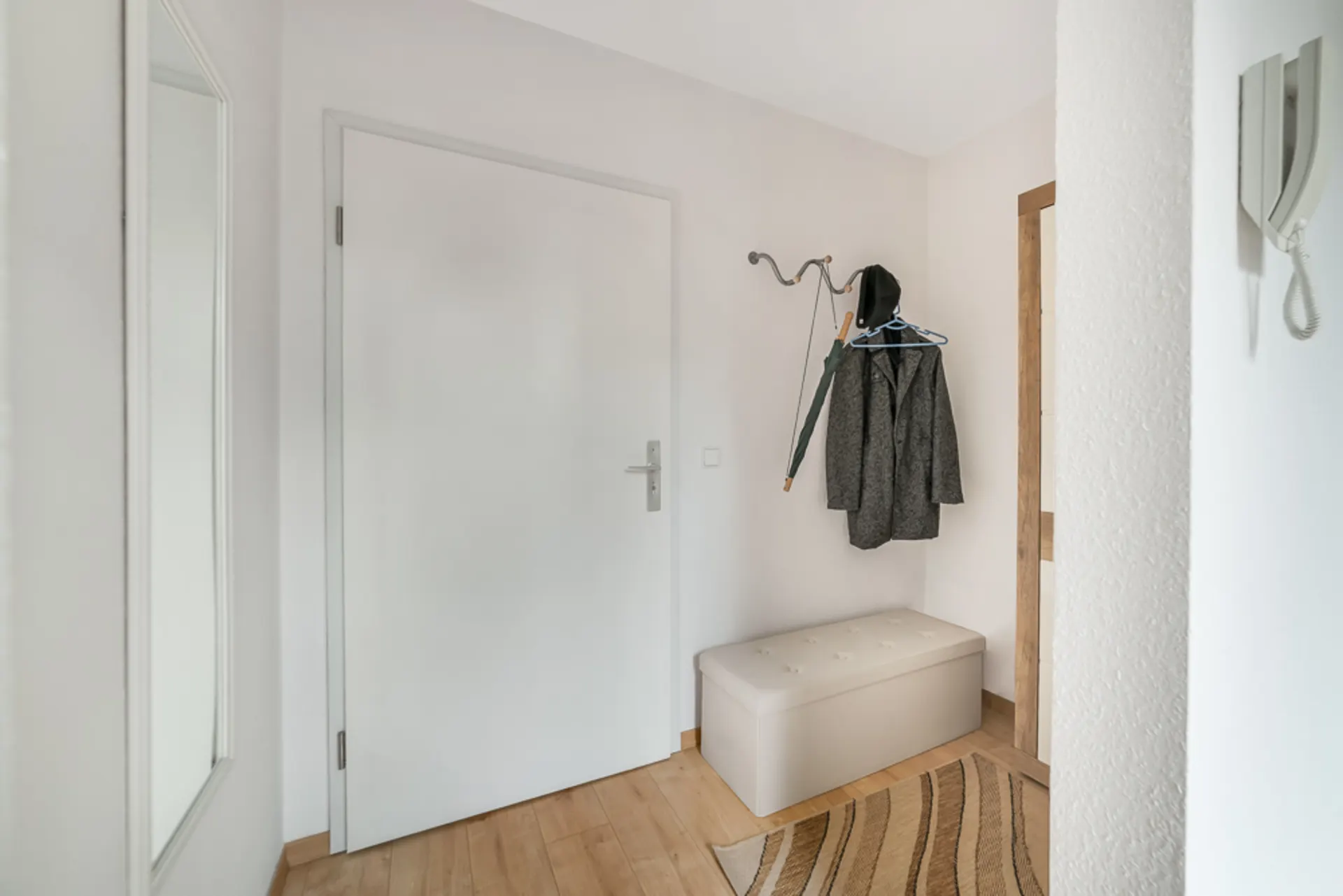 Heller Flur mit weißer Tür, Garderobe, Mantel, Regenschirm, Sitzbank und Spiegel.
