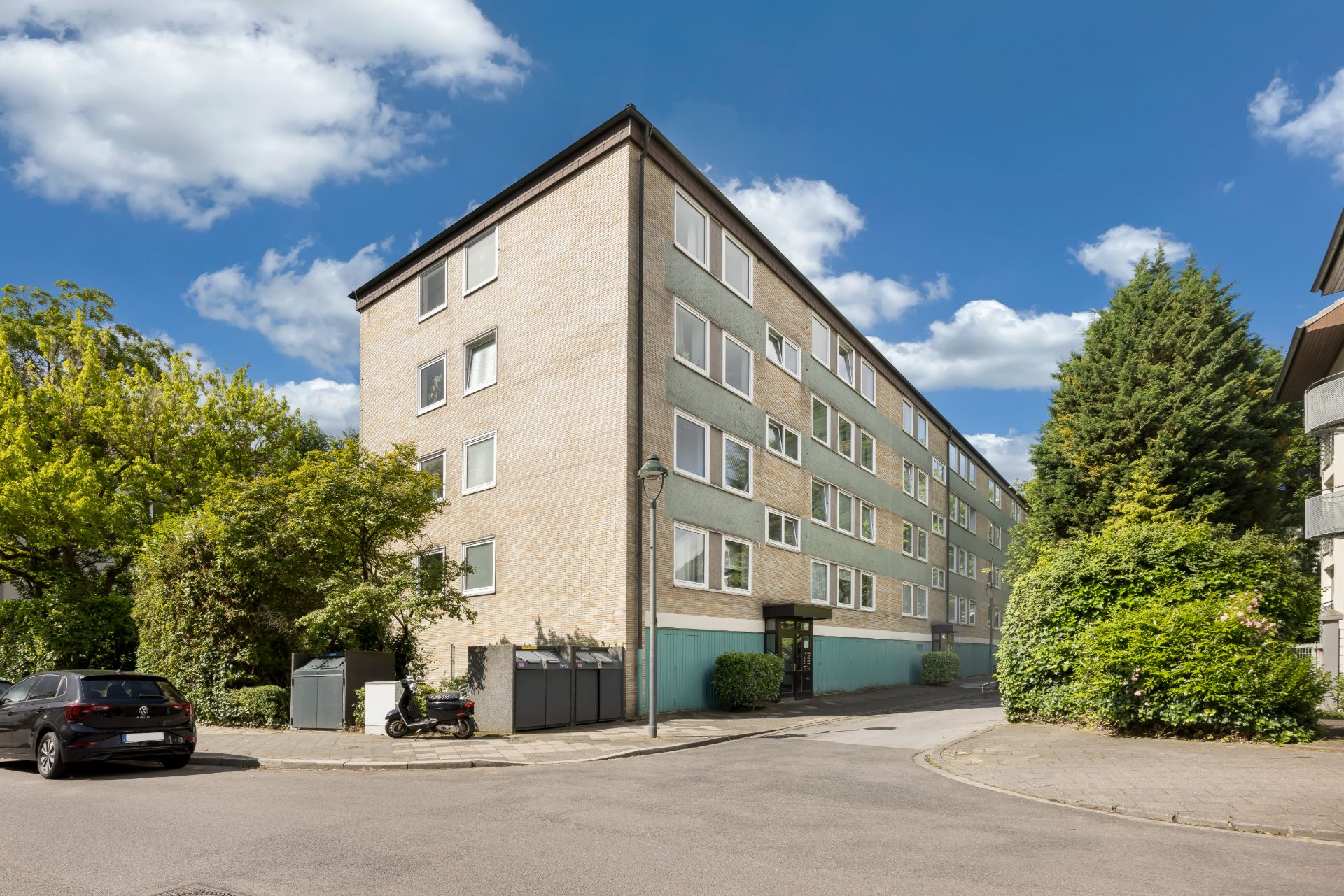 Mehrstöckiges Wohnhaus an einer Straßenecke mit Bäumen, Auto und Motorrad davor.