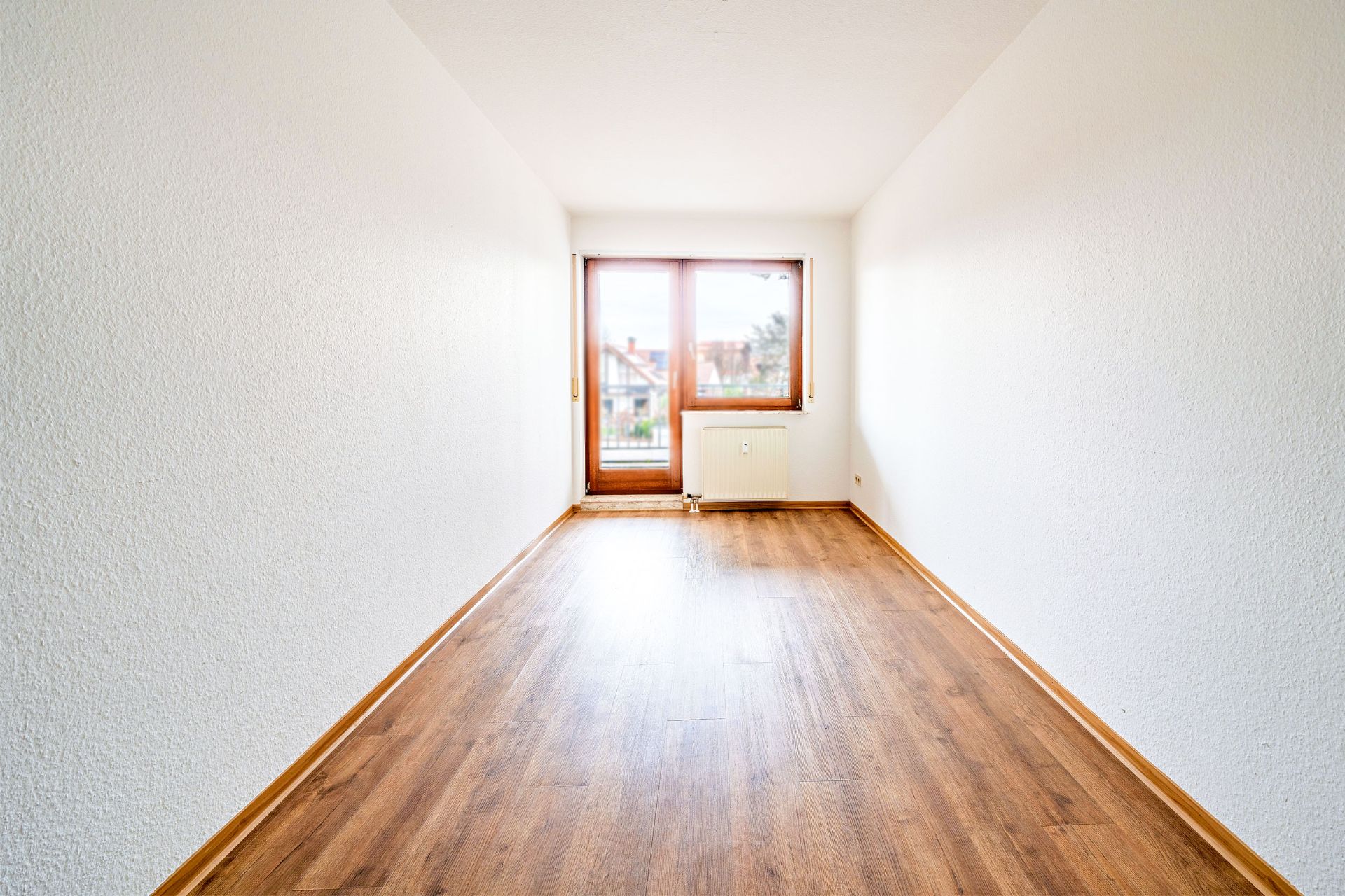 Leerer, heller Raum mit Holzboden und großem Fenster mit Tür zum Balkon.