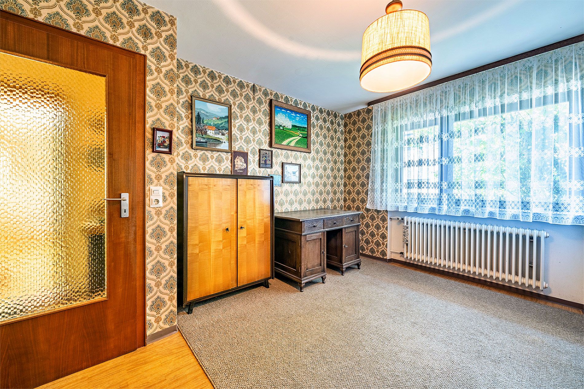 Heller Raum mit Holztür, Schrank, Schreibtisch, Teppichboden und großen Fenstern mit Gardinen.