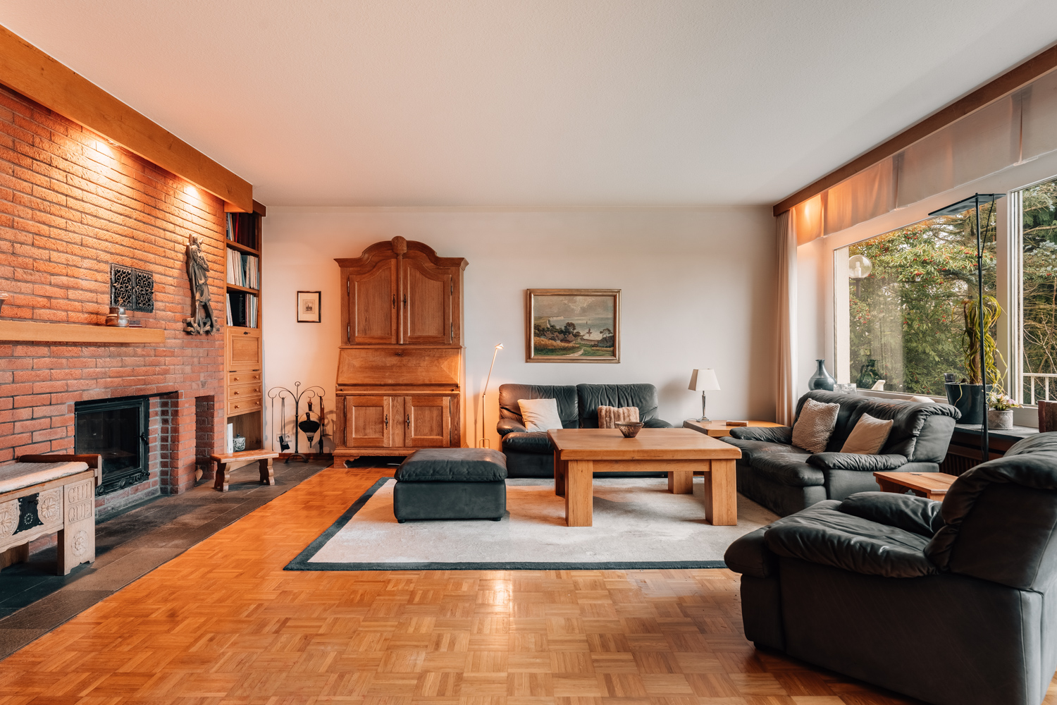 Gemütliches Wohnzimmer mit Kamin, Sofas, Holzmöbeln und großem Fenster mit Blick ins Grüne.