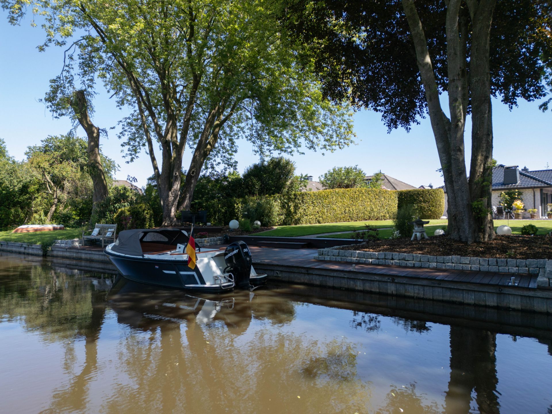 Kleines Boot am privaten Steg mit Garten, Bäumen und Haus im Hintergrund.