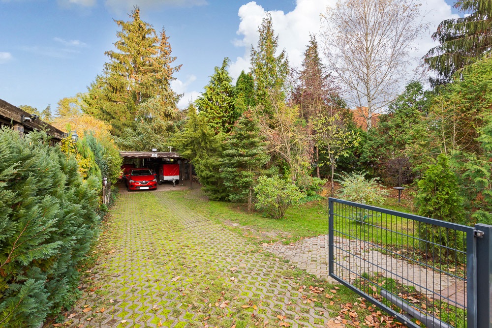 Einfahrt mit offenem Tor, gepflastertem Weg und Carport mit rotem Auto, umgeben von Bäumen.