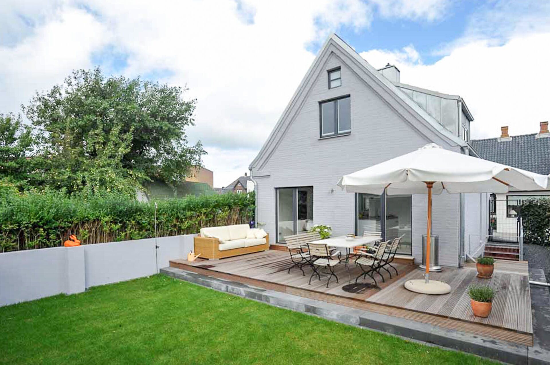 Modernes Haus mit Holzterrasse, Esstisch, Sonnenschirm und Sofa im Garten.