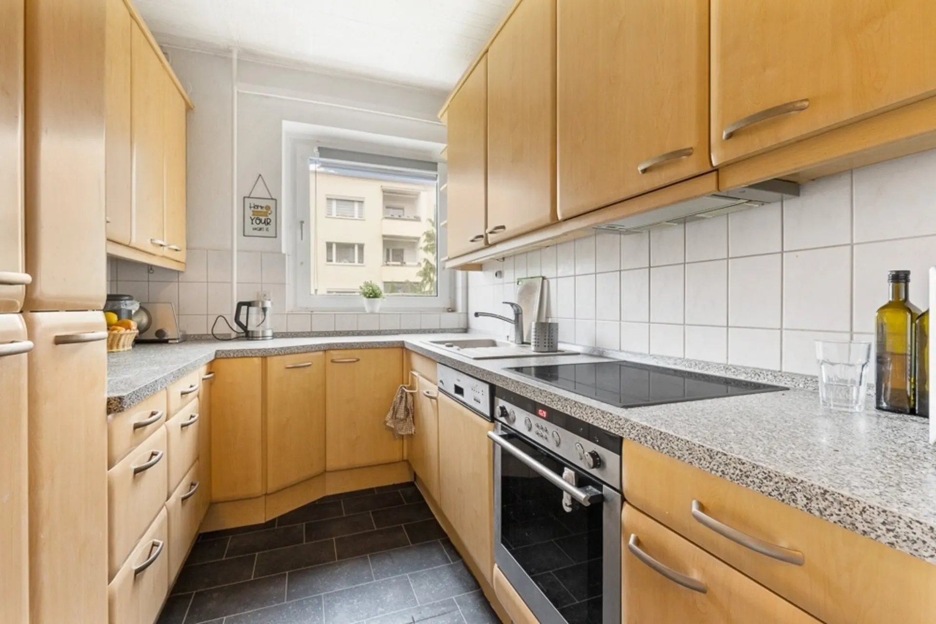 Moderne Küche mit hellen Holzschränken, Elektroherd, Spüle und Fenster mit Blick nach draußen.