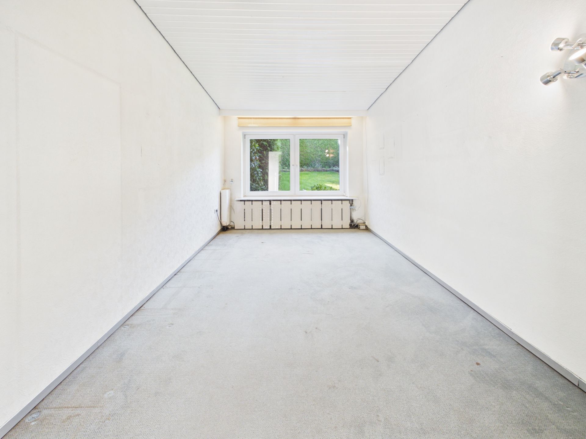 Leerer, schmaler Raum mit weißem Teppich und großem Fenster am Ende.