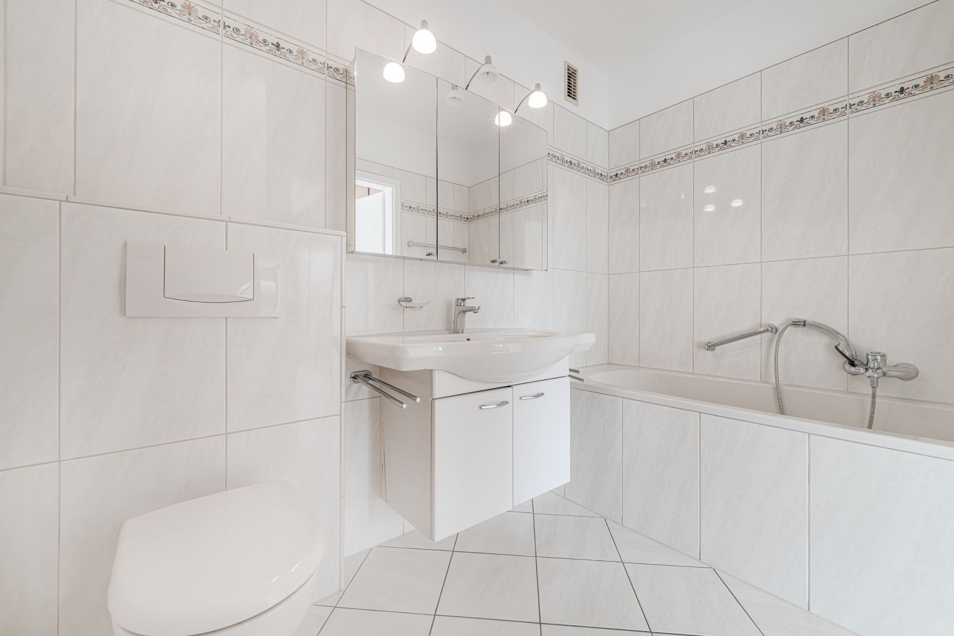 Helles Badezimmer mit Badewanne, Waschbecken, Spiegel und WC, alles in weiß gefliest.