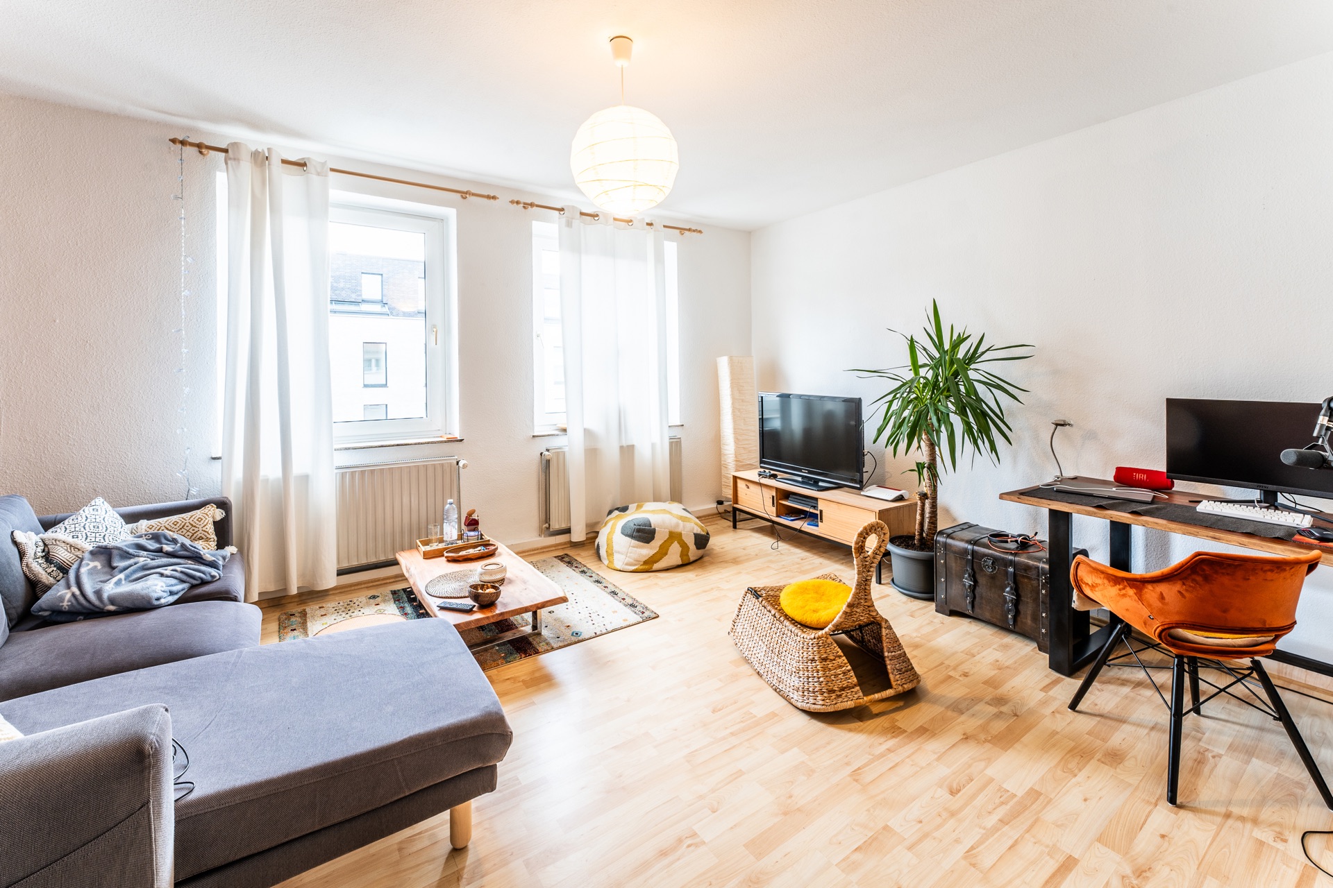 Helles Wohnzimmer mit Sofa, Couchtisch, Fernseher, Schreibtisch, Pflanze und zwei Fenstern.