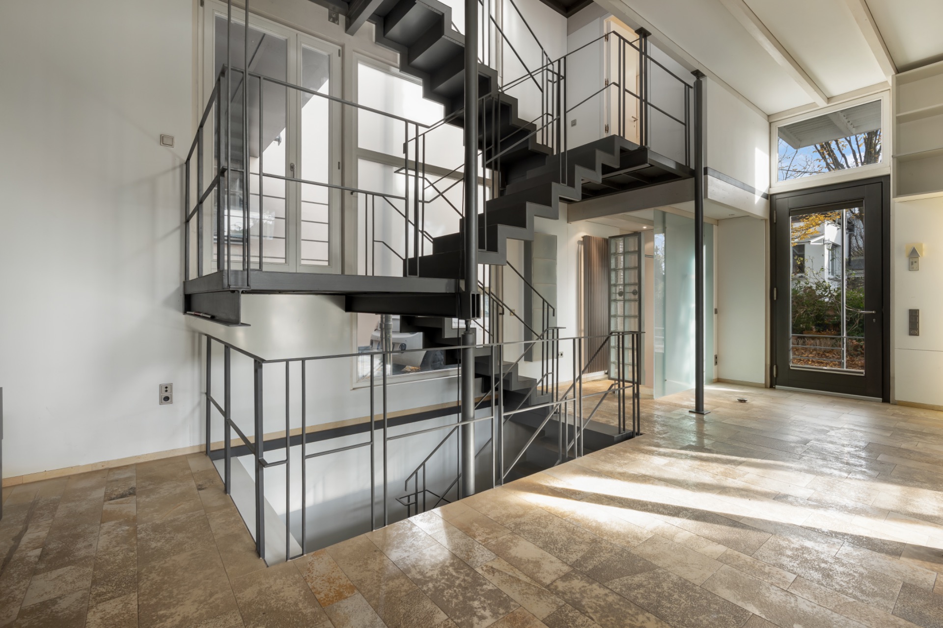 Heller Eingangsbereich mit Metalltreppe und großen Fenstern, Steinboden.
