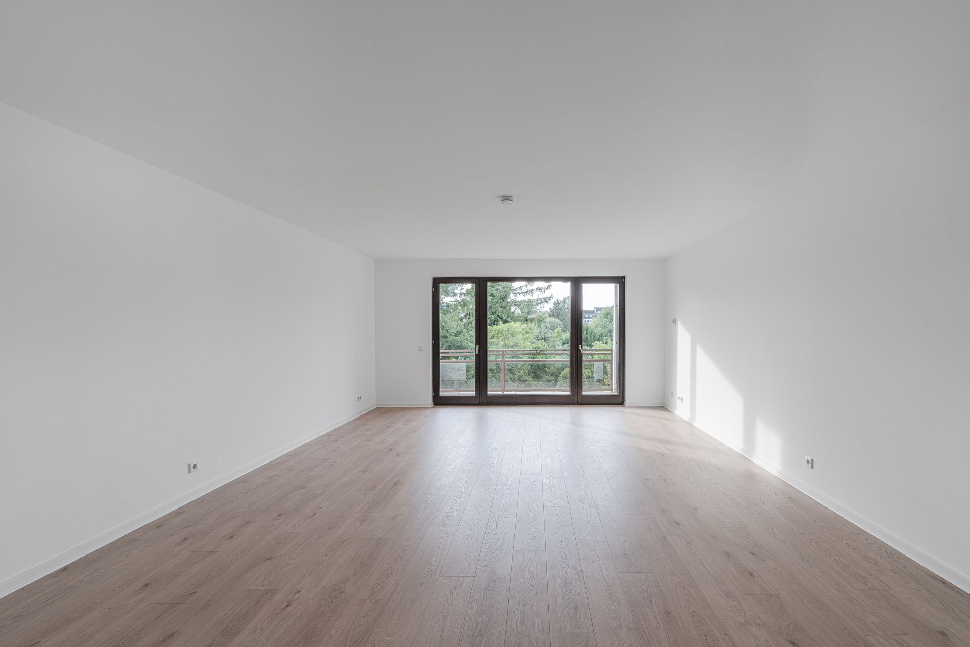 Leerer, heller Raum mit Holzfußboden und großen Fenstern mit Blick ins Grüne.