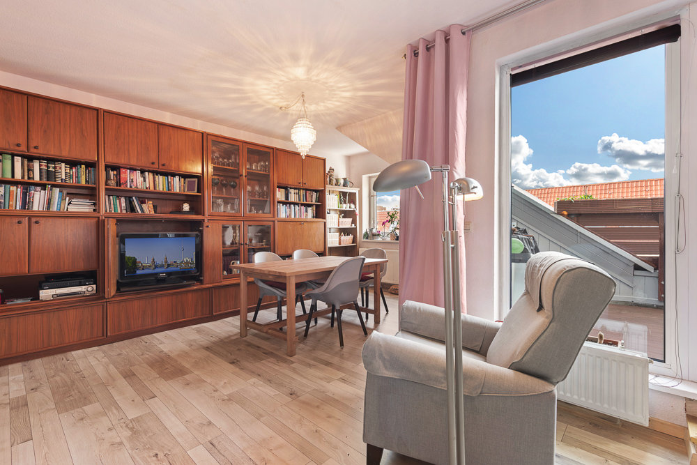 Wohnzimmer mit Holzregal, Sessel, Esstisch und Zugang zu einem Balkon.
