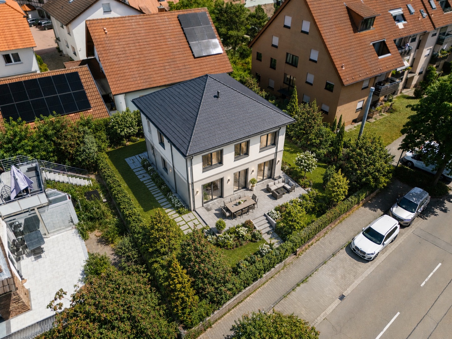 Luftaufnahme eines modernen Einfamilienhauses mit Garten und Terrasse an einer Wohnstraße.