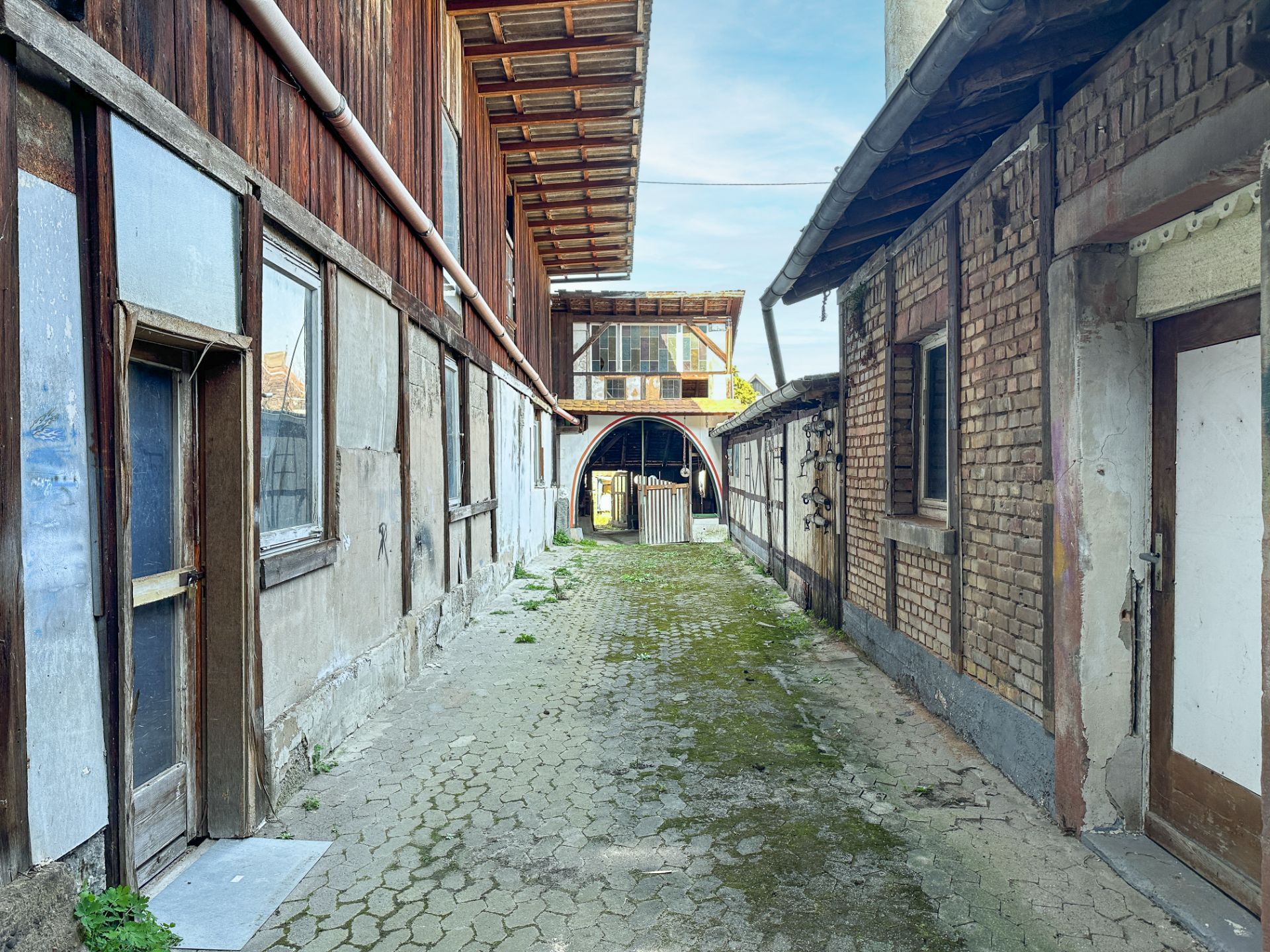 Gepflasterter Hof mit zwei alten Gebäuden, sichtbaren Türen und Fenstern, überdachter Durchgang im Hintergrund.