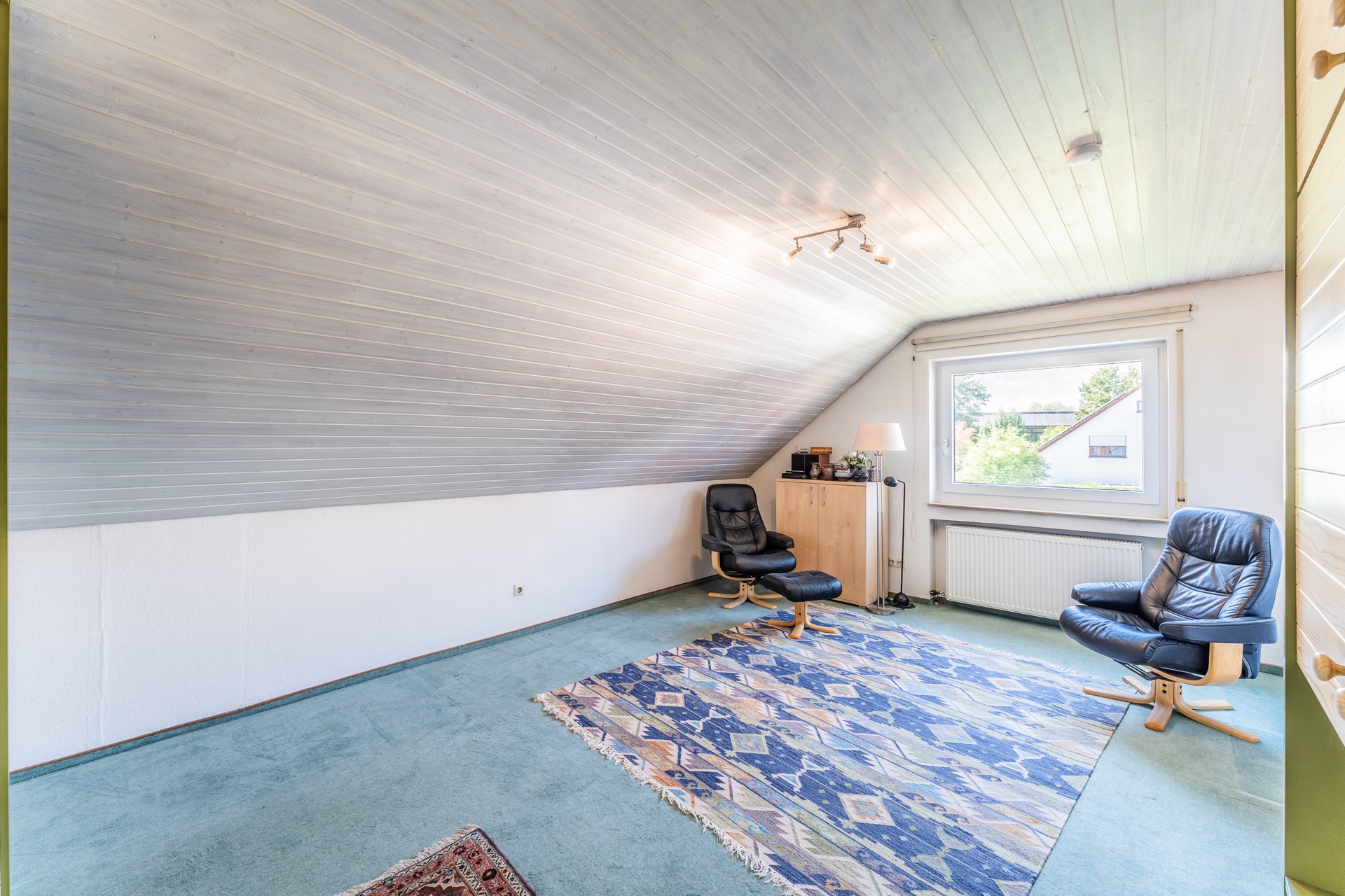 Helles Zimmer mit schräger Holzdecke, zwei Sesseln, Teppich und großem Fenster.