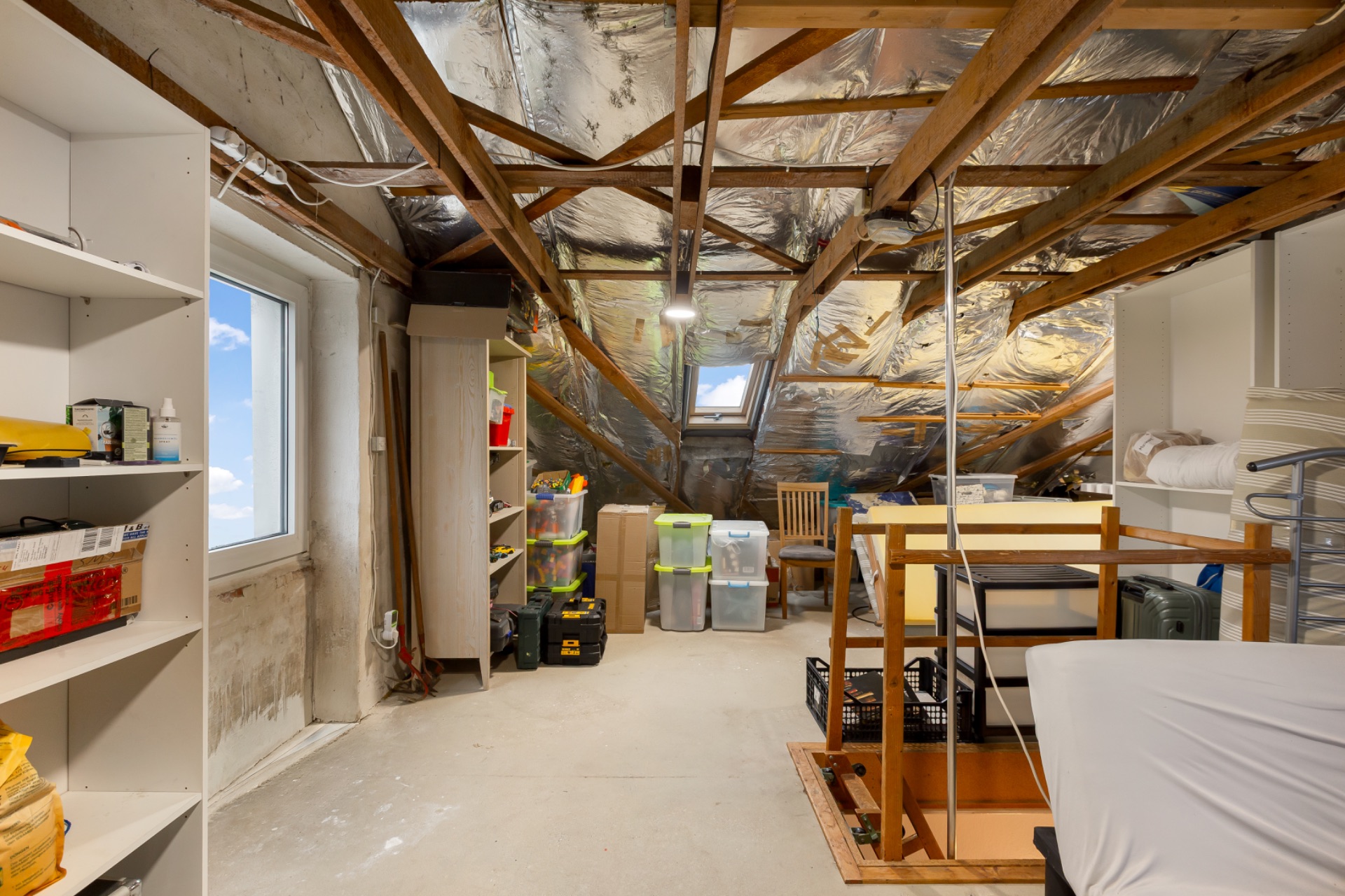 Dachboden mit Holzträgern, Lageregale, Kisten und einem Fenster.