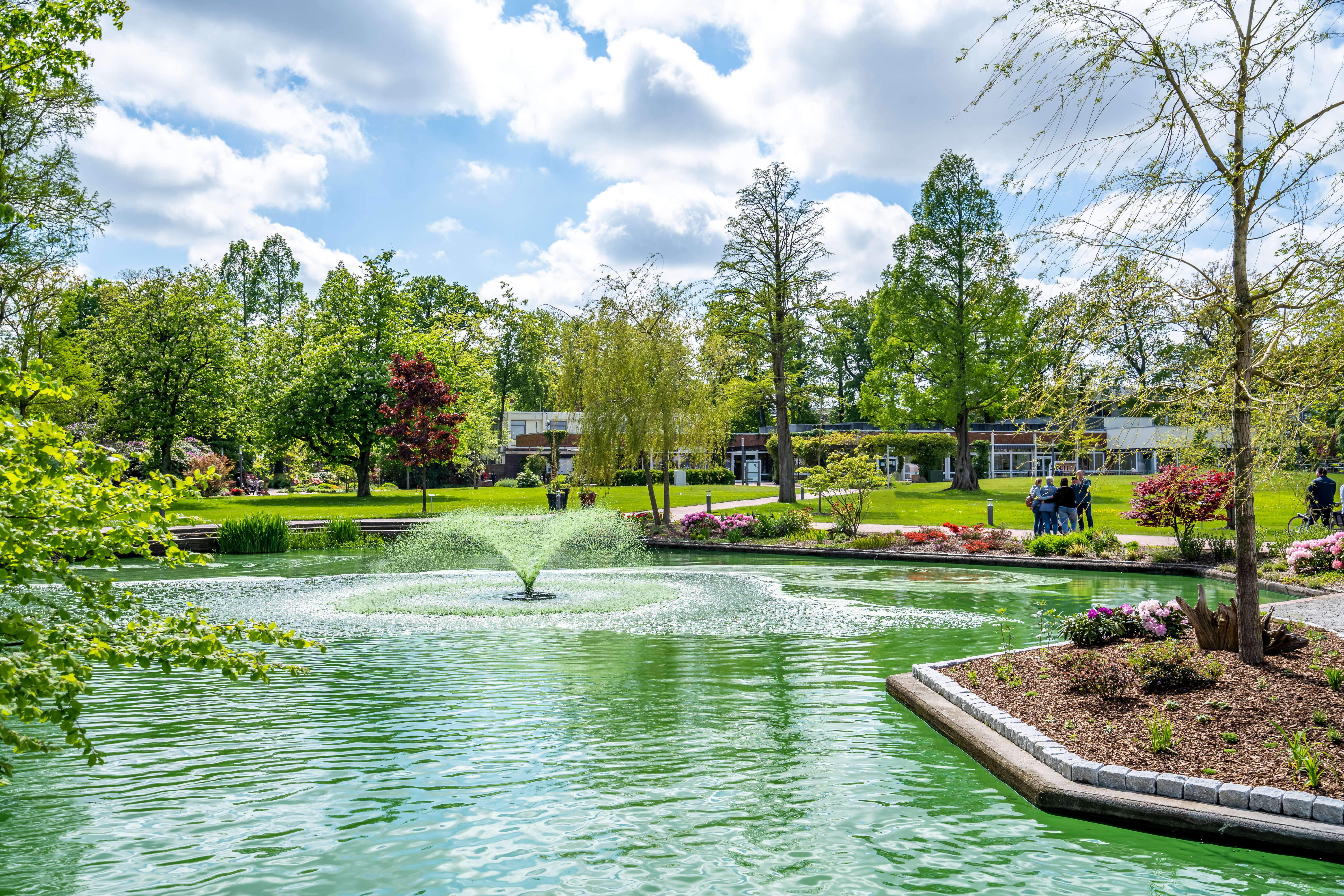 Garten mit kleinem Teich, Springbrunnen und Blumenbeeten, im Hintergrund Bäume und Gebäude.