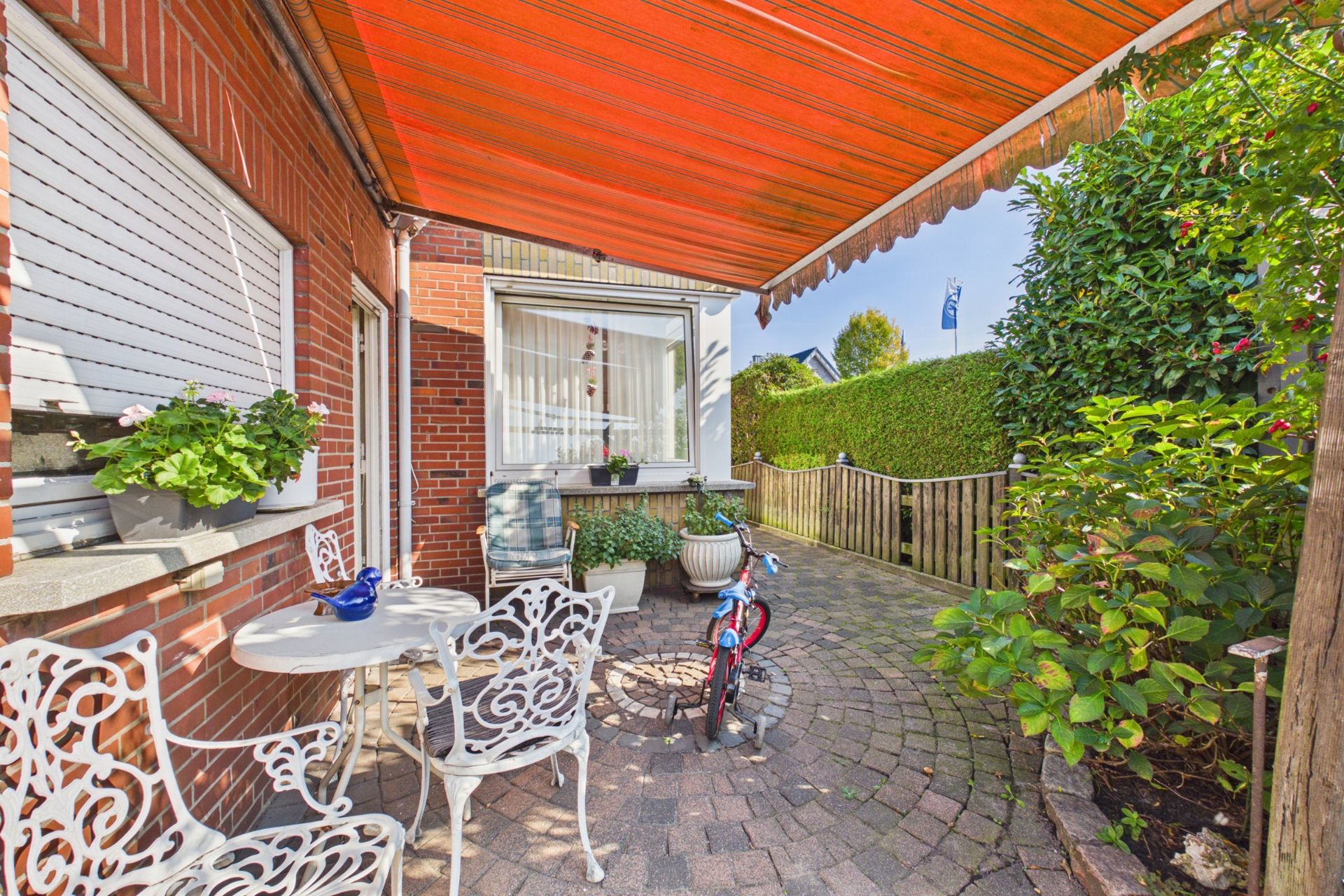 Gepflasterte Terrasse mit weißen Gartenmöbeln, Kinderfahrrad und orangefarbener Markise.
