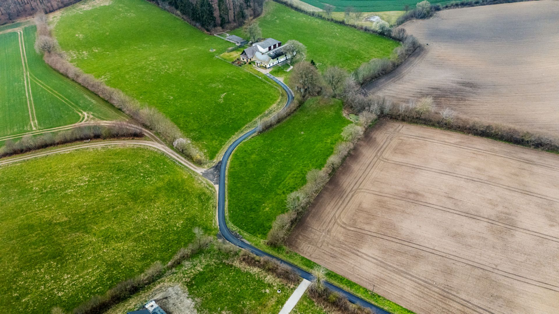 Luftaufnahme eines Hauses auf einem großen, grünen Grundstück mit umliegenden Feldern und Zufahrtsstraße.
