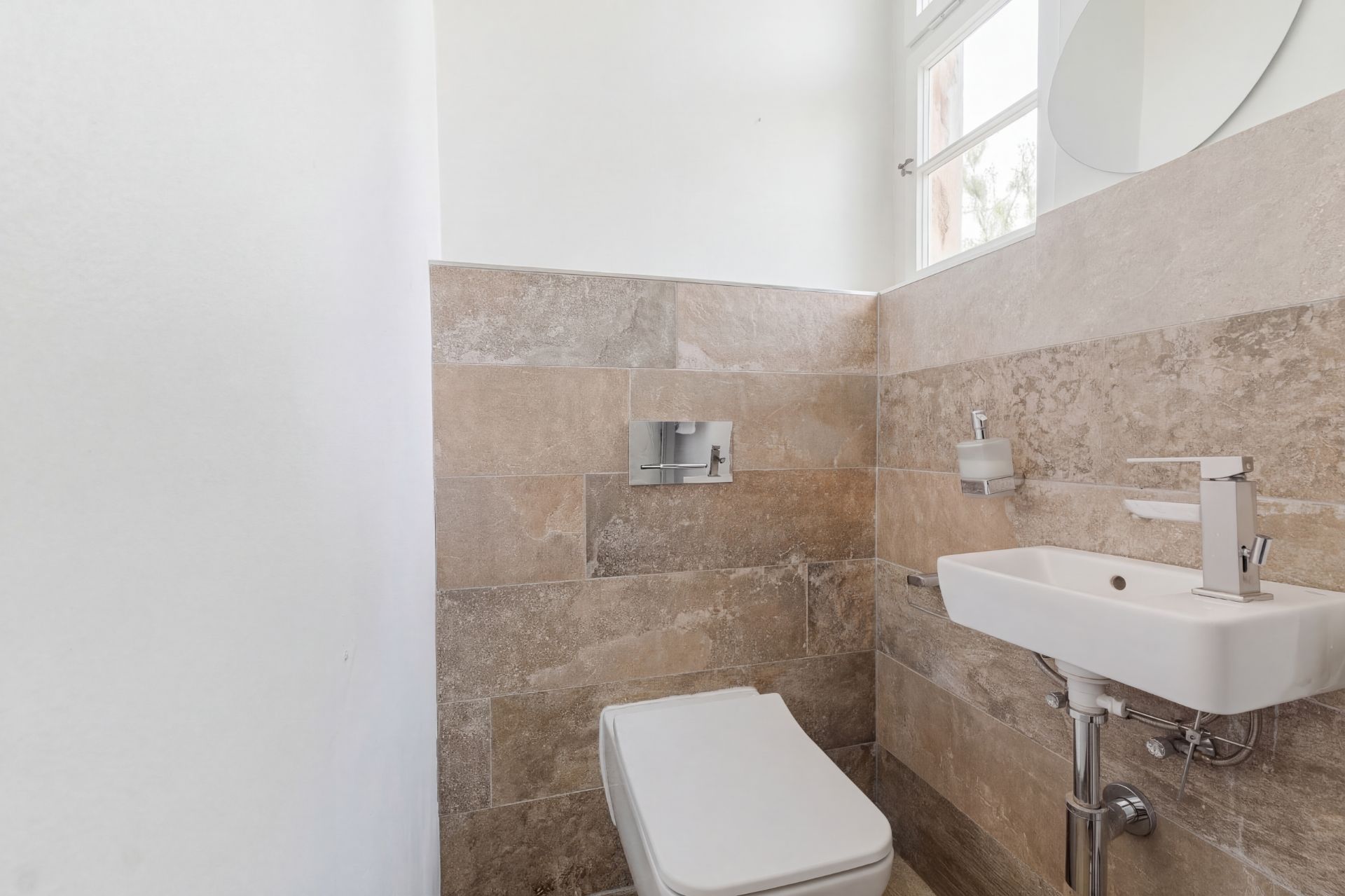 Kleines Badezimmer mit WC, Waschbecken und Fenster, Wände mit beigen Fliesen.