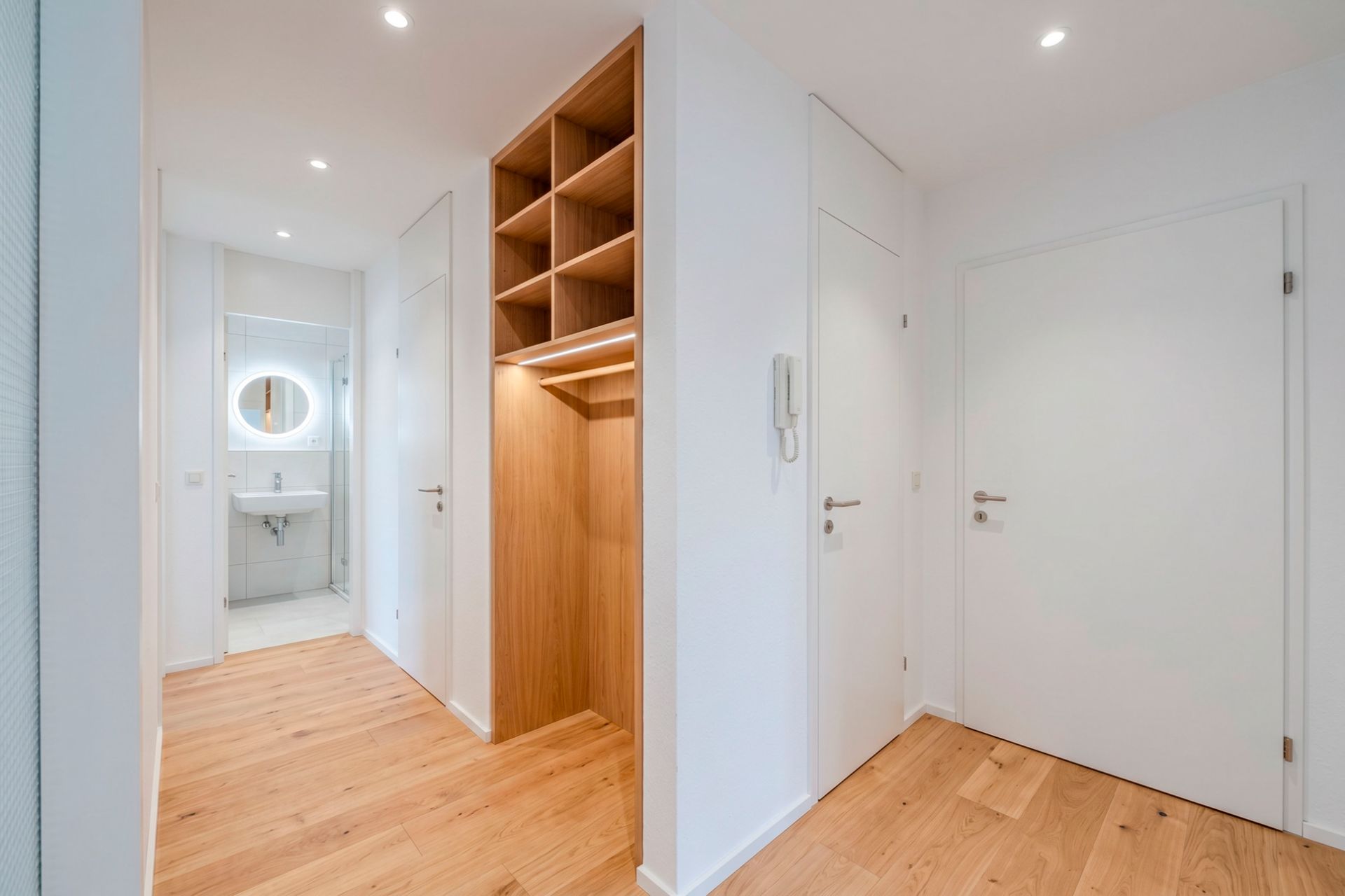 Heller Flur mit Holzregal, Parkettboden und Blick ins Badezimmer mit rundem Spiegel.