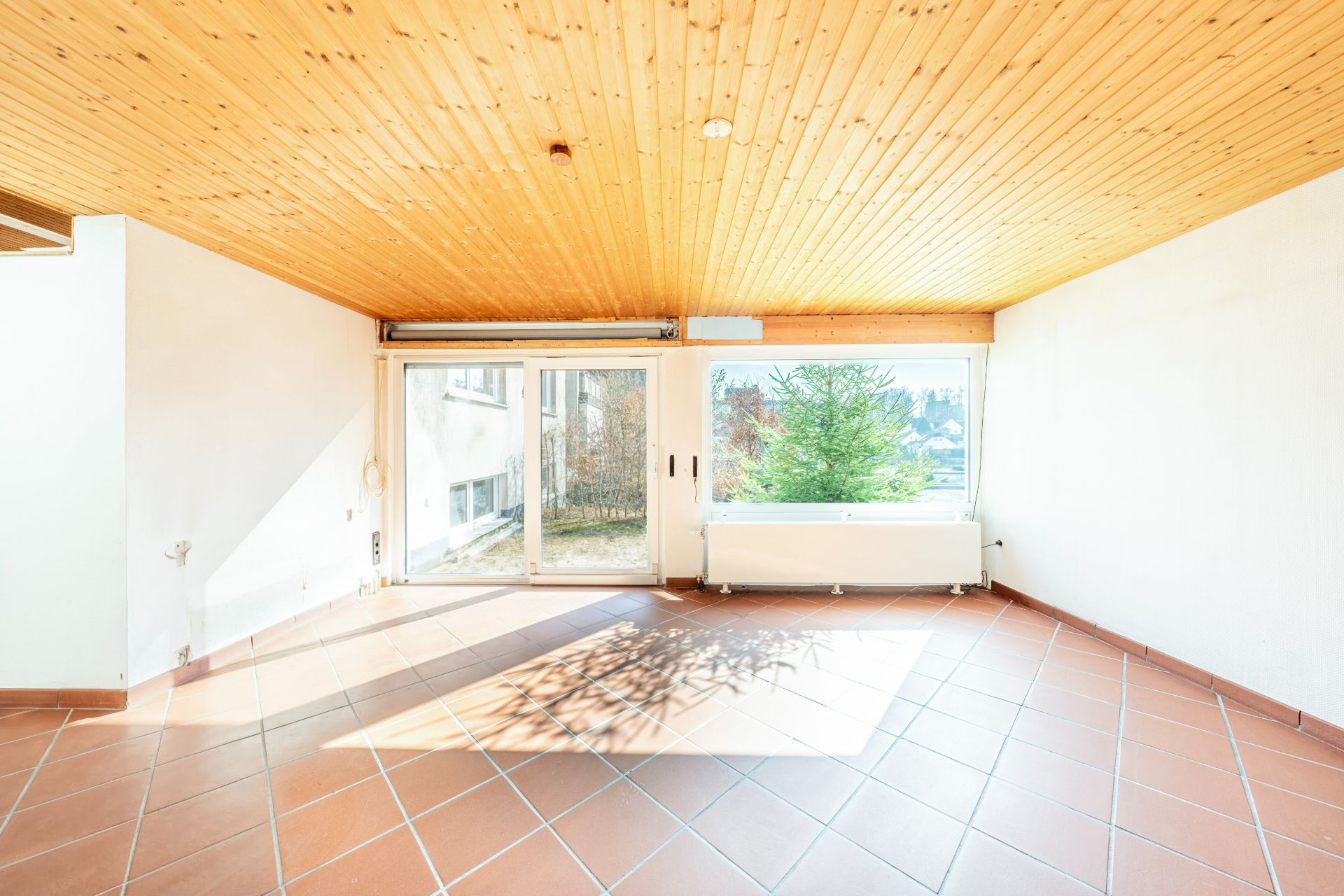 Heller Raum mit Holzdecke, Fliesenboden und großen Fenstern zur Terrasse mit Blick ins Grüne.