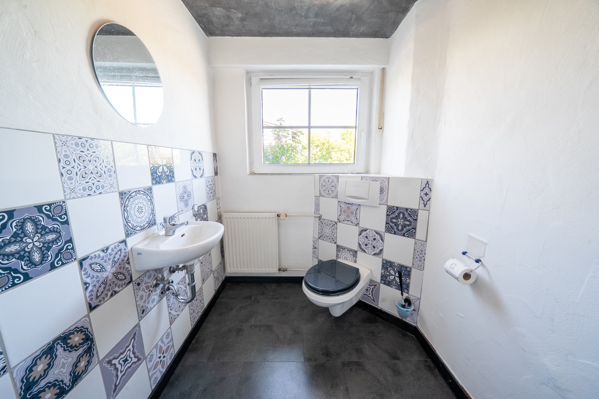 Helles Badezimmer mit gemusterten Fliesen, Wandwaschbecken, WC und Fenster.