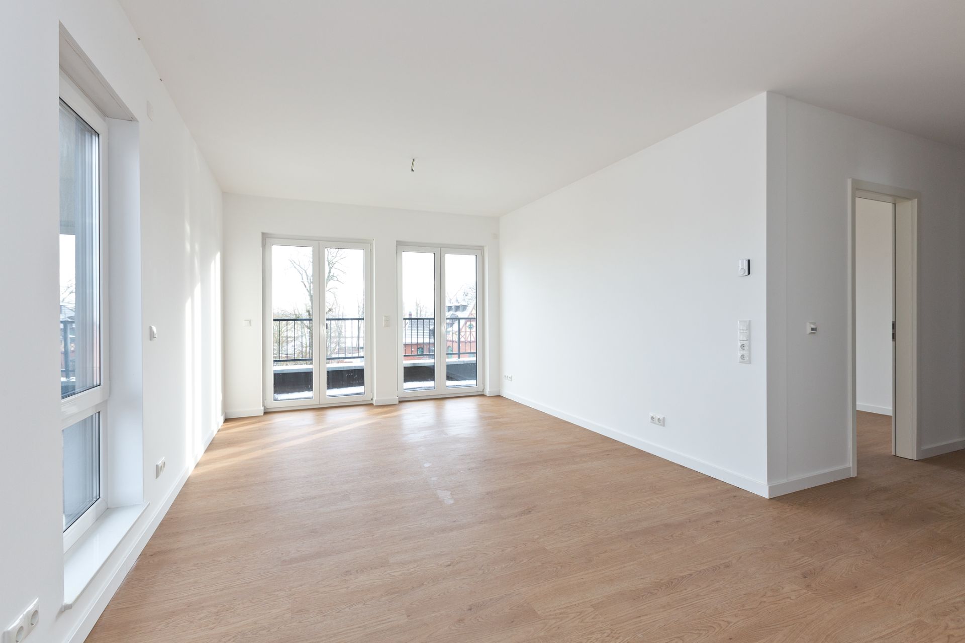 Helles Wohnzimmer mit Holzboden, großen Fenstern und Zugang zu einem Balkon.
