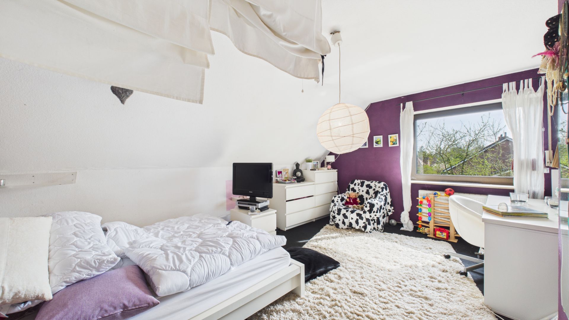 Helles Schlafzimmer mit Bett, Kommode, Sessel, Teppich und Schreibtisch am Fenster.