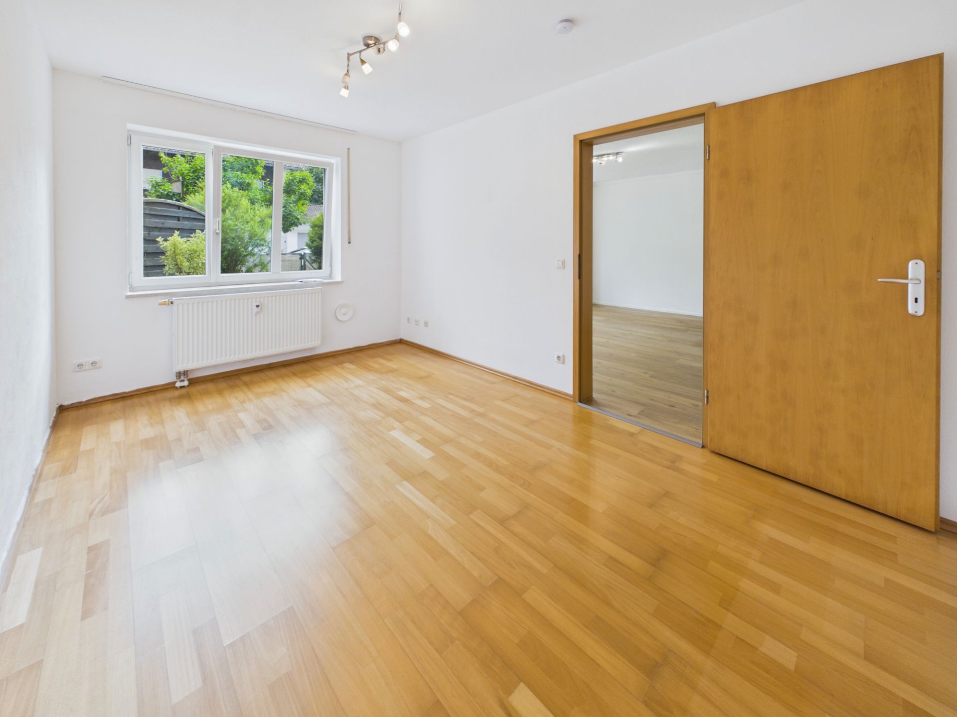 Helles Zimmer mit Parkettboden, großem Fenster und geöffneter Holztür zu einem zweiten Raum.