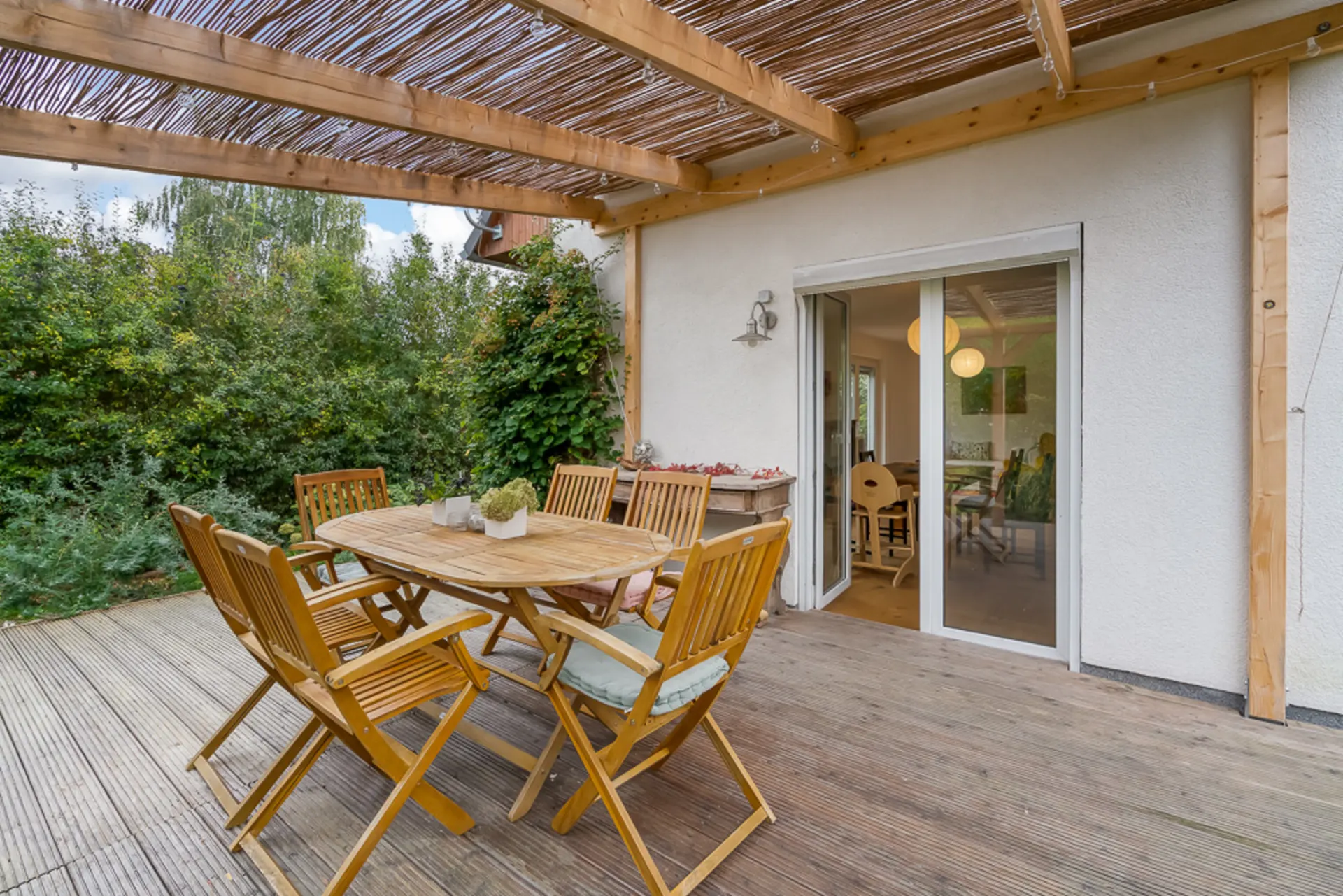 Überdachte Terrasse mit Holztisch und sechs Stühlen, Schiebetür zum Innenraum, grüne Hecke im Hintergrund.